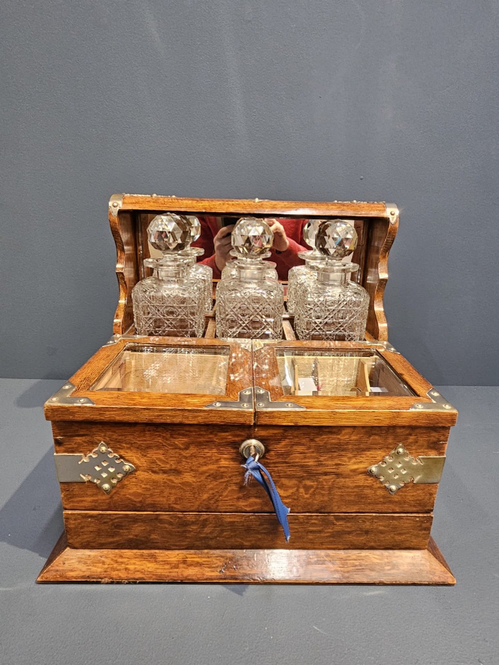 A Victorian oak tantalus featuring brass corner mounts, three cut-glass decanters with faceted stoppers set against a mirrored back, and forward-folding compartments for gaming accessories. View IV