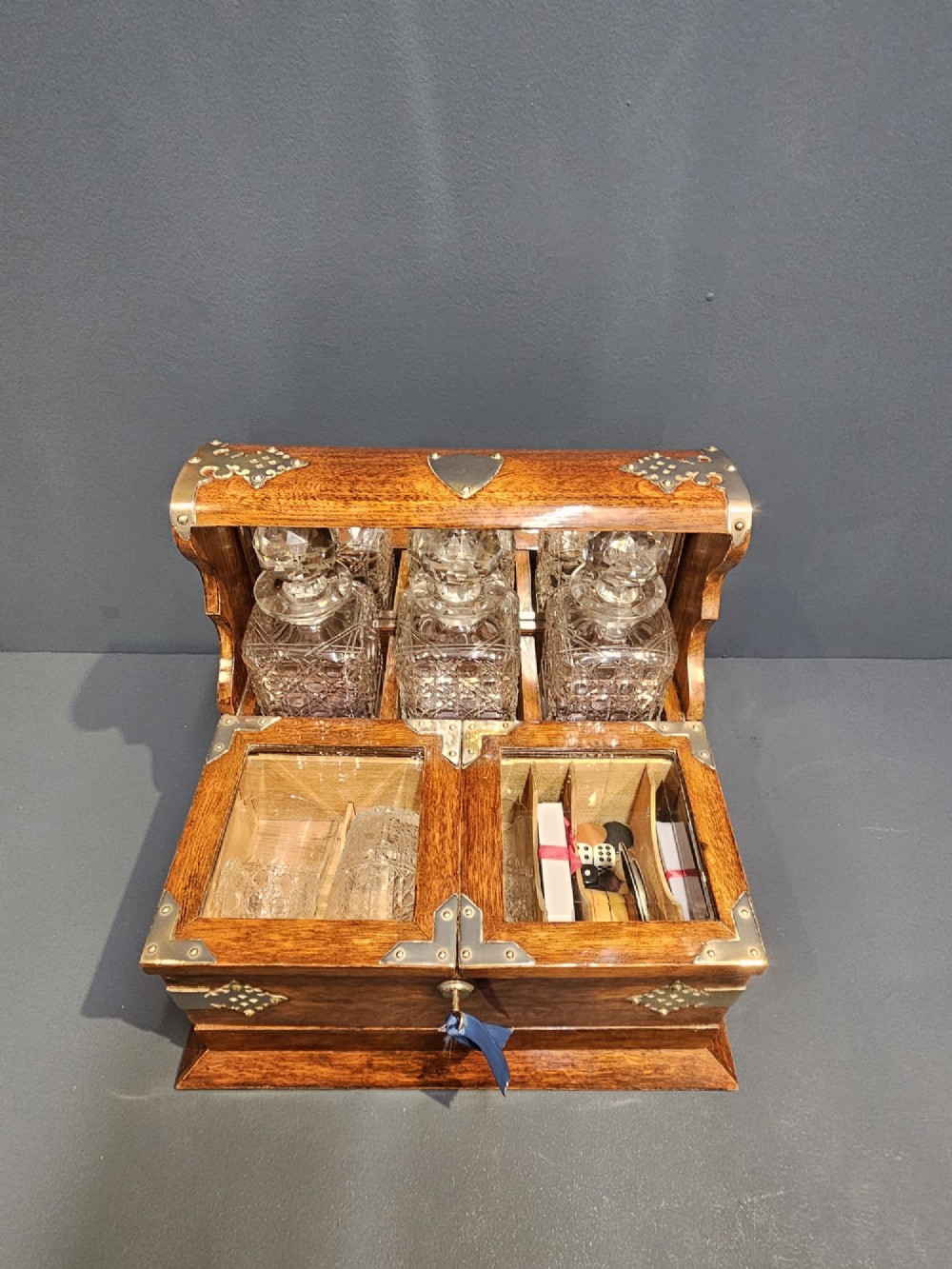 A Victorian oak tantalus featuring brass corner mounts, three cut-glass decanters with faceted stoppers set against a mirrored back, and forward-folding compartments for gaming accessories. View V