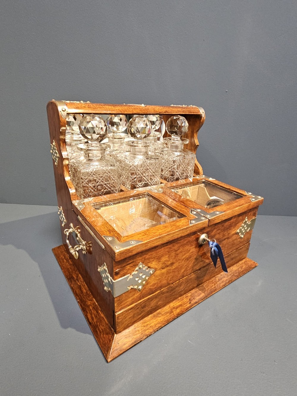 A Victorian oak tantalus featuring brass corner mounts, three cut-glass decanters with faceted stoppers set against a mirrored back, and forward-folding compartments for gaming accessories. View VI