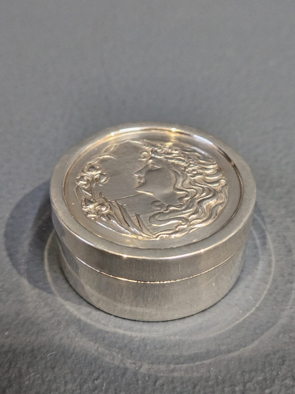 A circular silver pill box featuring an embossed Art Nouveau profile of a woman with flowing hair and flora on the top lid.