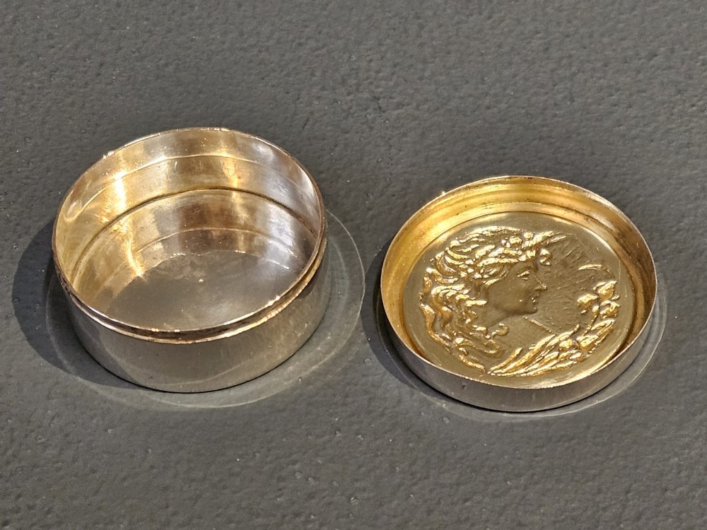 A circular silver pill box featuring an embossed Art Nouveau profile of a woman with flowing hair and flora on the top lid. View II
