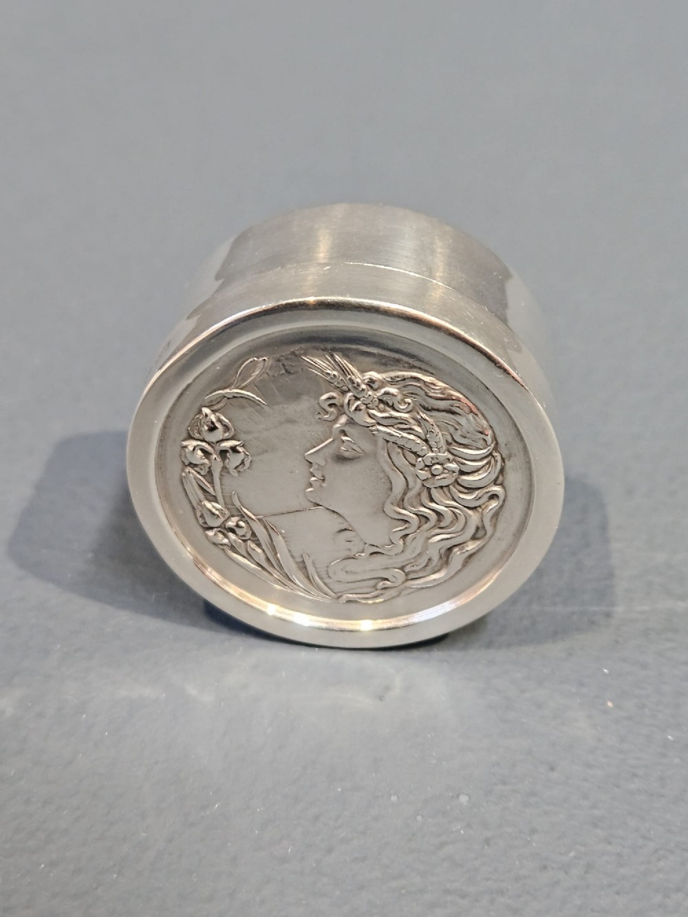 A circular silver pill box featuring an embossed Art Nouveau profile of a woman with flowing hair and flora on the top lid. View IV