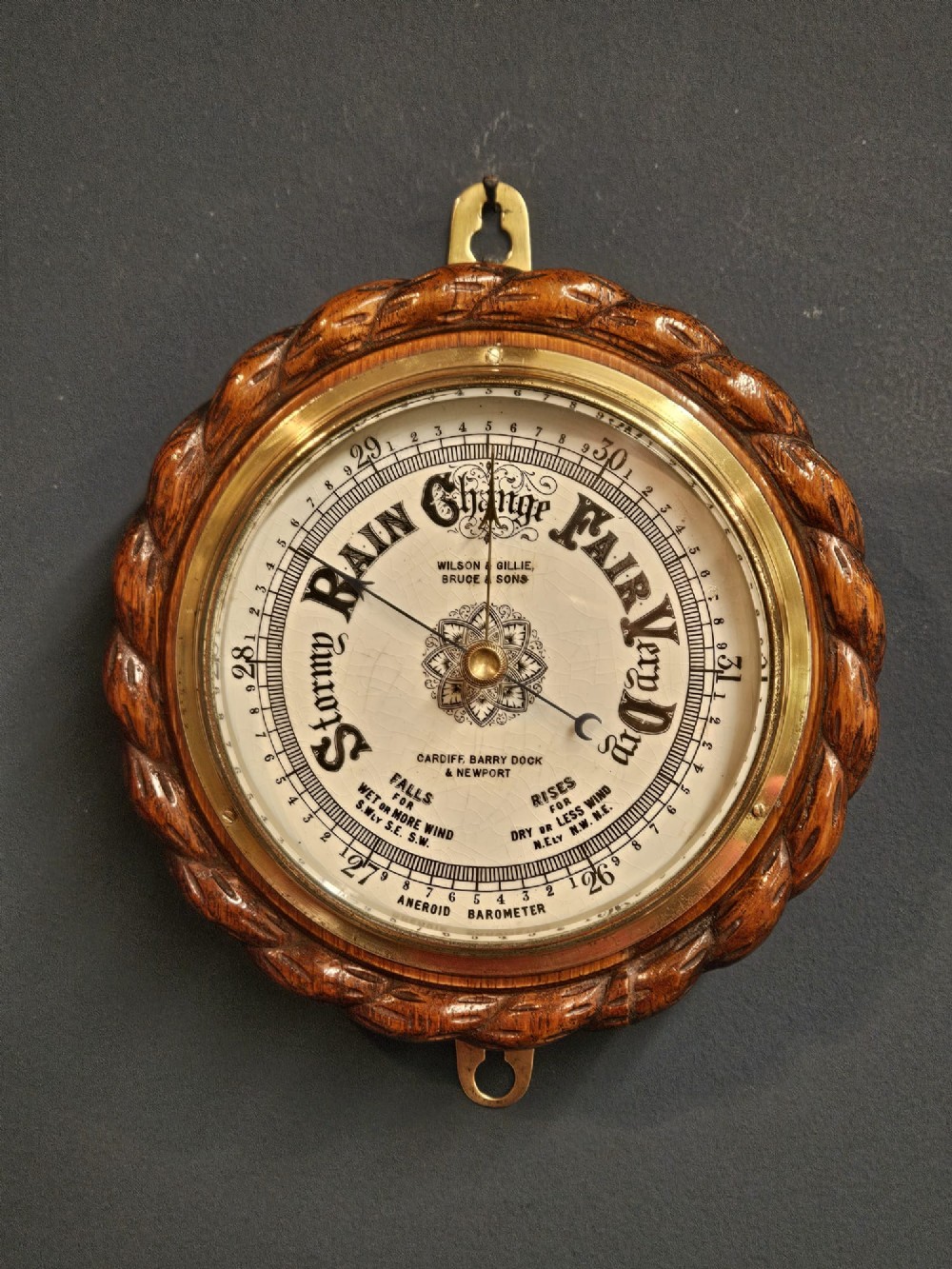 A circular antique oak aneroid barometer featuring a carved rope twist border, brass bezel, and detailed ceramic dial with black lettering.