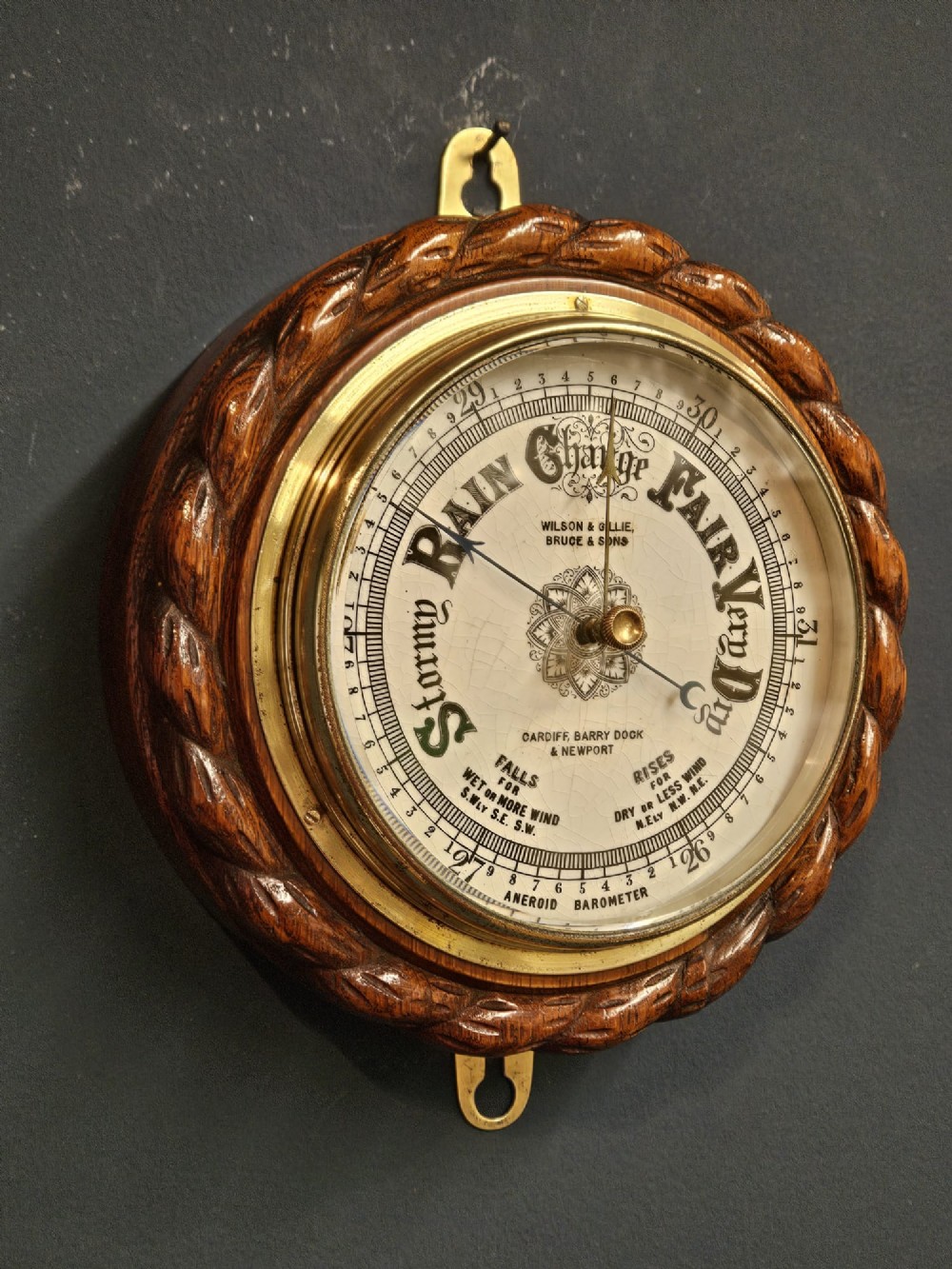 A circular antique oak aneroid barometer featuring a carved rope twist border, brass bezel, and detailed ceramic dial with black lettering. View II