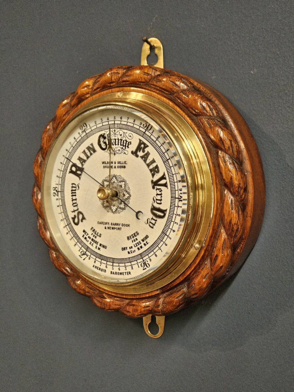 A circular antique oak aneroid barometer featuring a carved rope twist border, brass bezel, and detailed ceramic dial with black lettering. View III