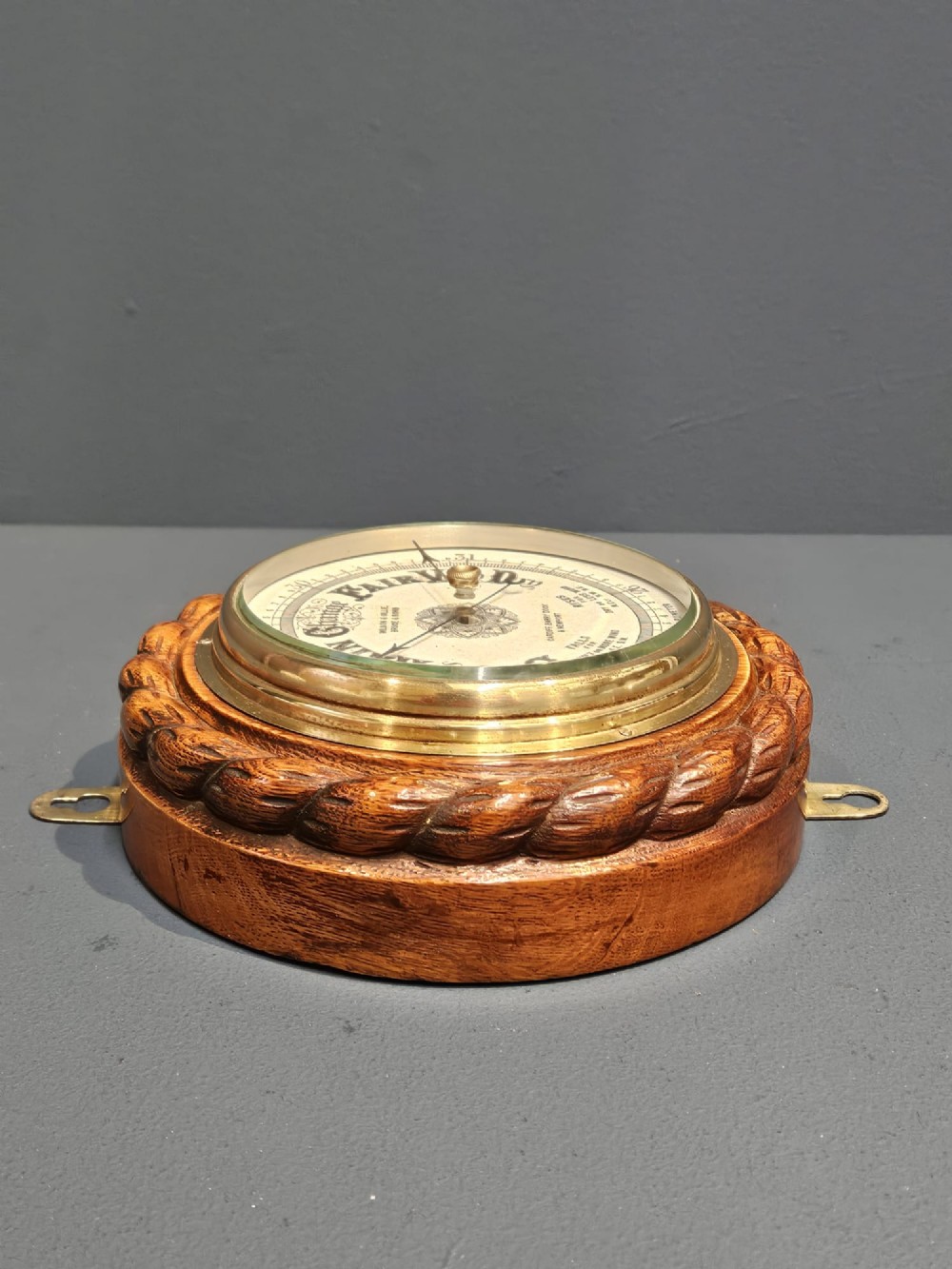 A circular antique oak aneroid barometer featuring a carved rope twist border, brass bezel, and detailed ceramic dial with black lettering. View V