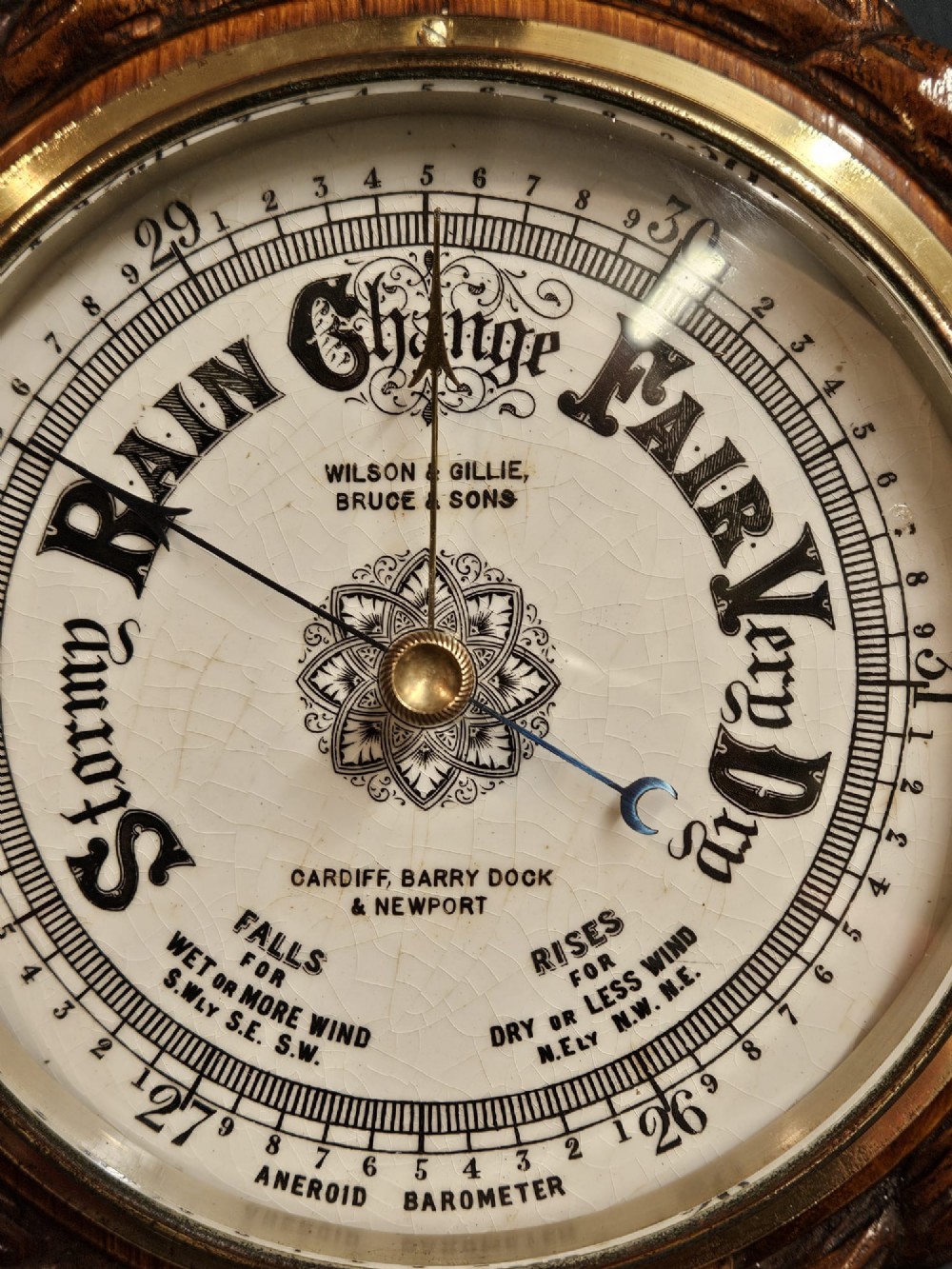 A circular antique oak aneroid barometer featuring a carved rope twist border, brass bezel, and detailed ceramic dial with black lettering. View VI