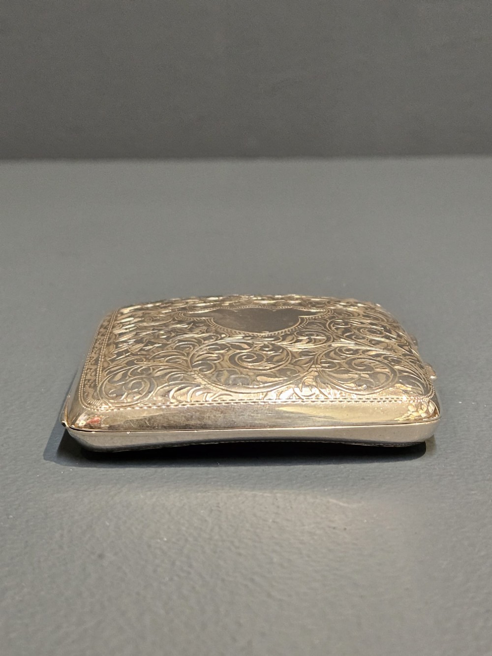 A square metallic silver pocket case featuring detailed chased foliate scrollwork and a central blank cartouche. View V