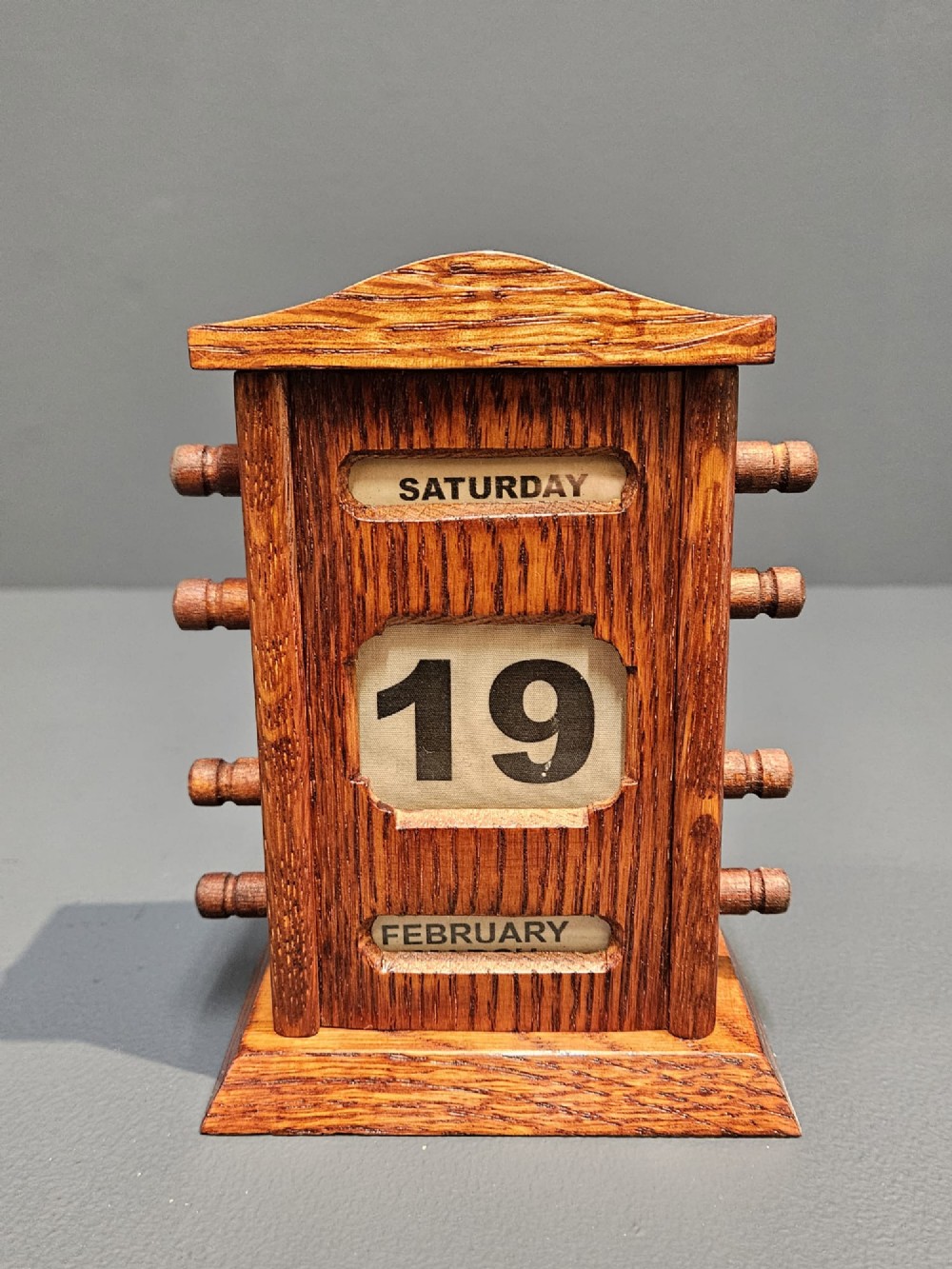 An Edwardian carved oak perpetual desk calendar featuring side turning pegs and printed linen date scrolls behind rectangular window openings.