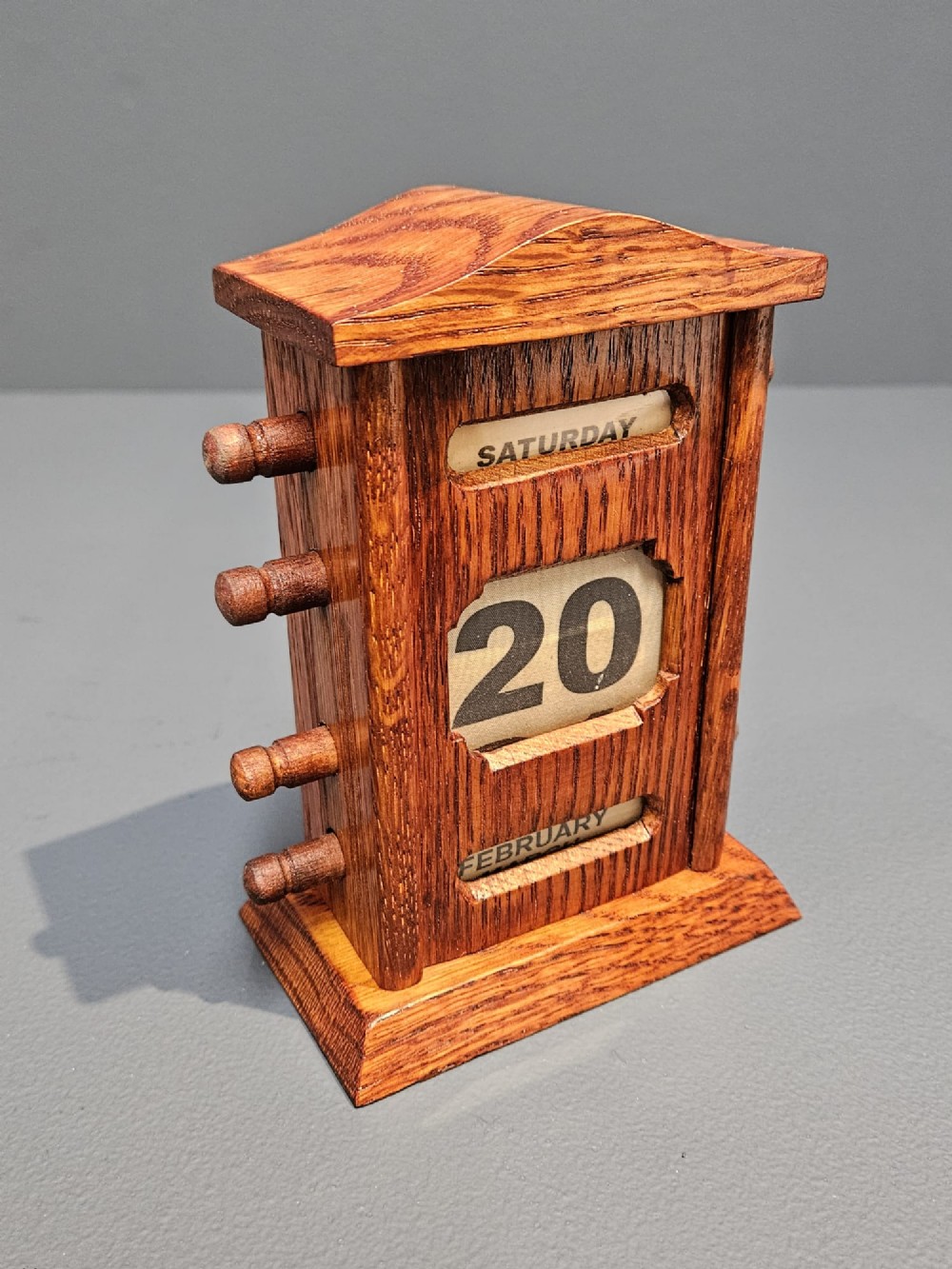 An Edwardian carved oak perpetual desk calendar featuring side turning pegs and printed linen date scrolls behind rectangular window openings. View II