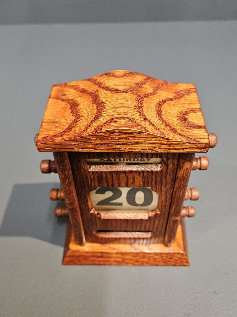 An Edwardian carved oak perpetual desk calendar featuring side turning pegs and printed linen date scrolls behind rectangular window openings. View III