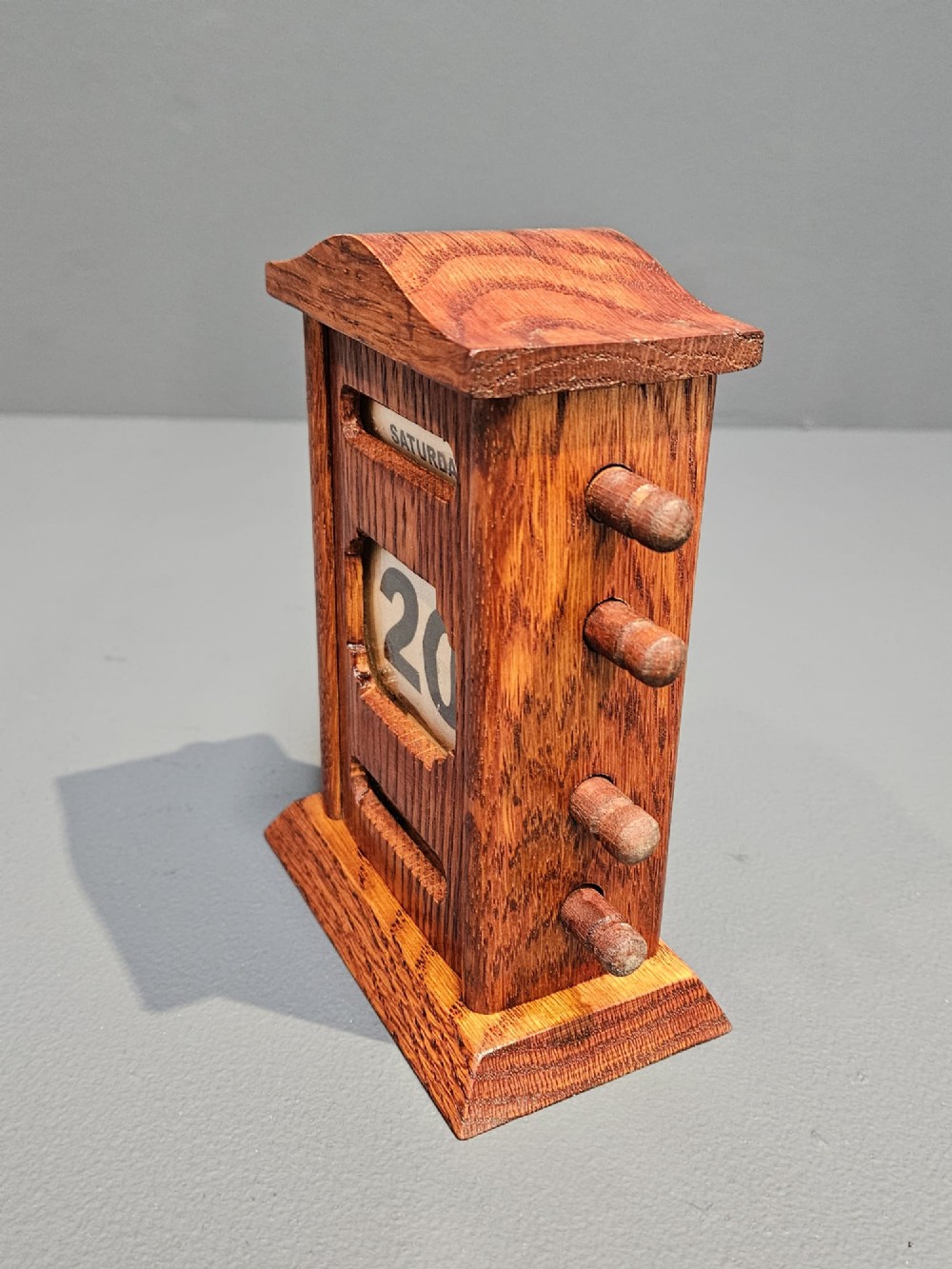 An Edwardian carved oak perpetual desk calendar featuring side turning pegs and printed linen date scrolls behind rectangular window openings. View VI