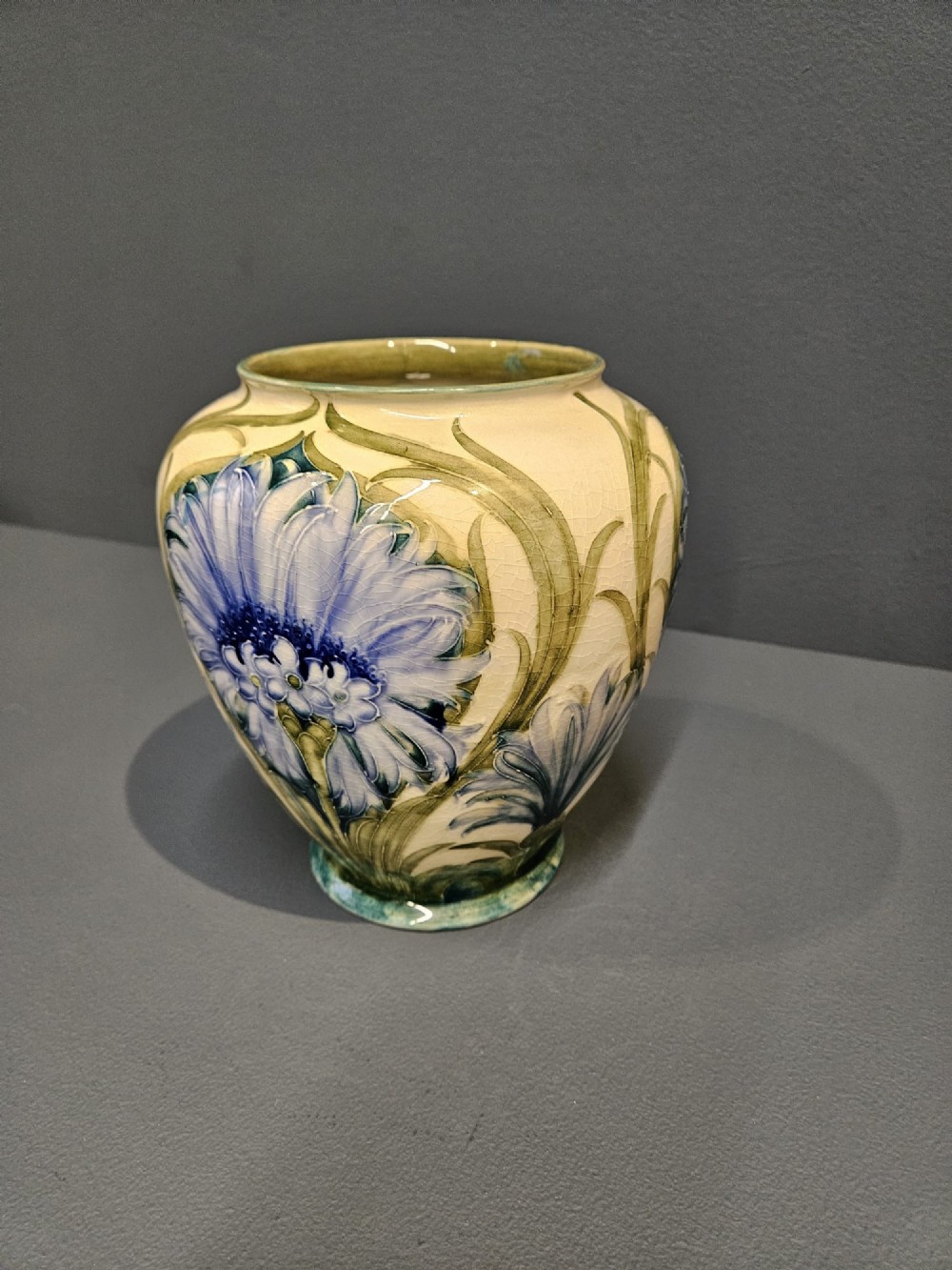 A bulbous ceramic vase featuring tube-lined blue cornflower motifs with green foliage set against a cream background. View VI