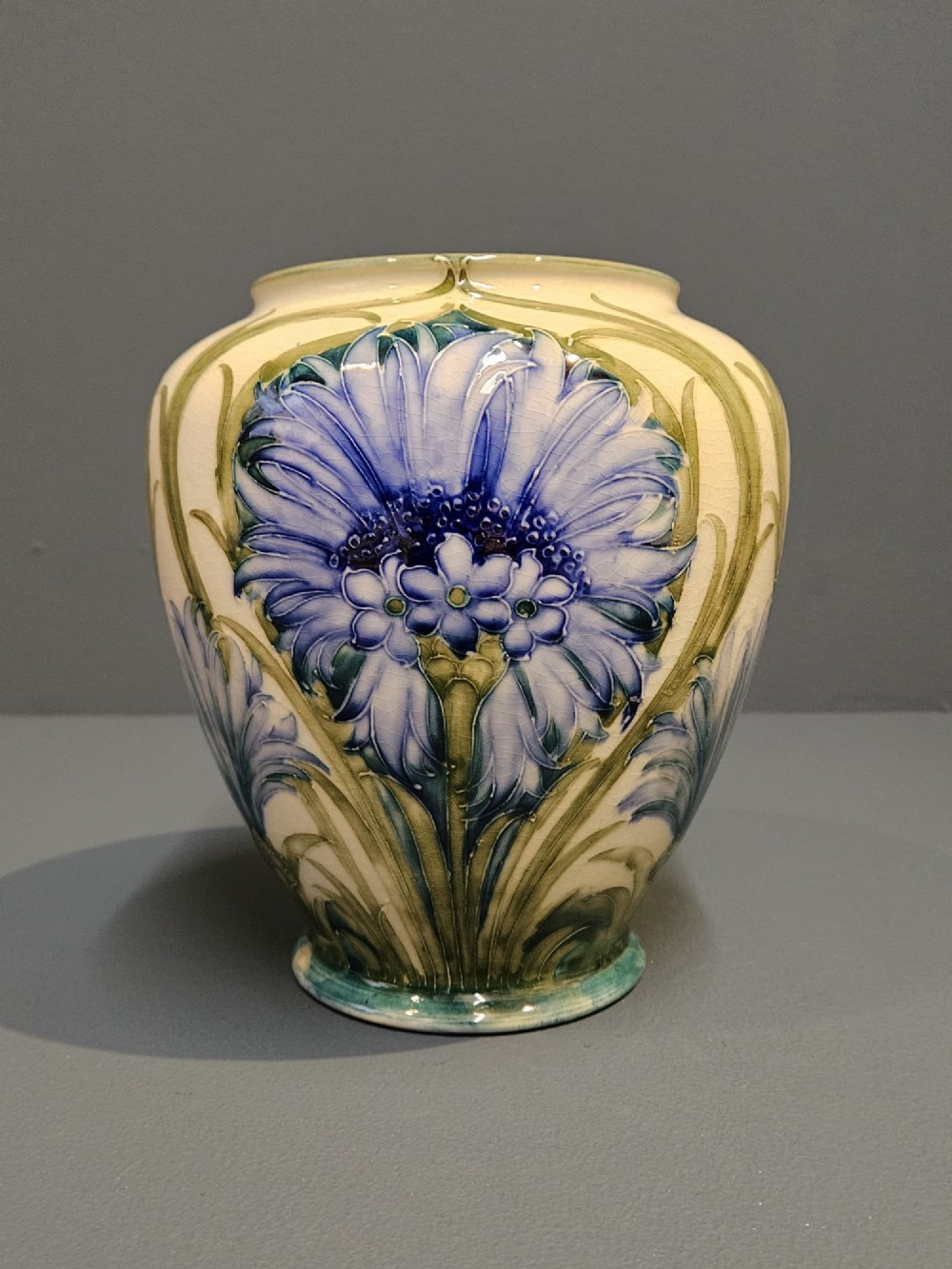 A bulbous ceramic vase featuring tube-lined blue cornflower motifs with green foliage set against a cream background. View VII