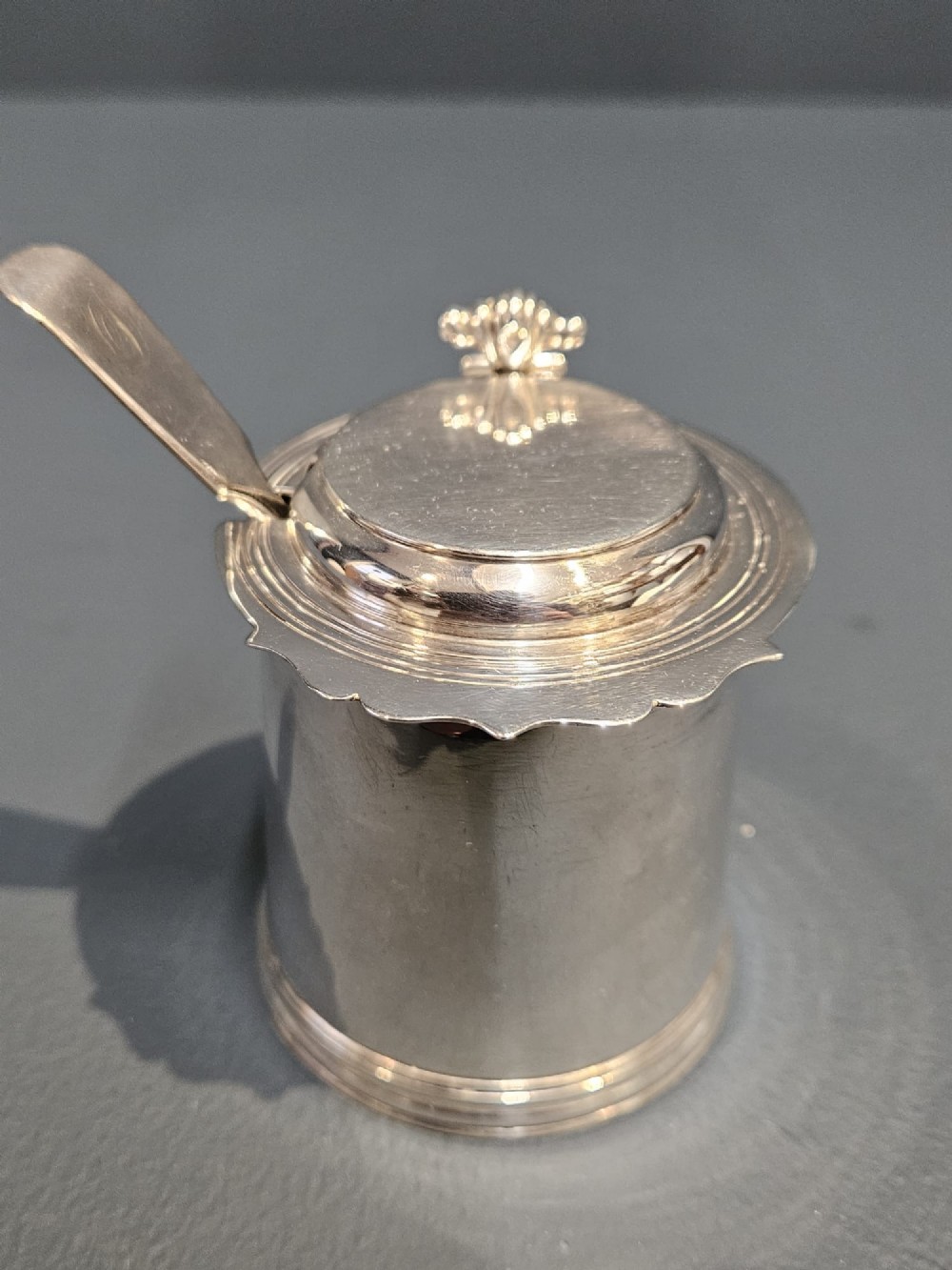 A cylindrical Britannia silver mustard pot featuring a scalloped collar rim, stepped lid with shell finial, and an inserted silver spoon.