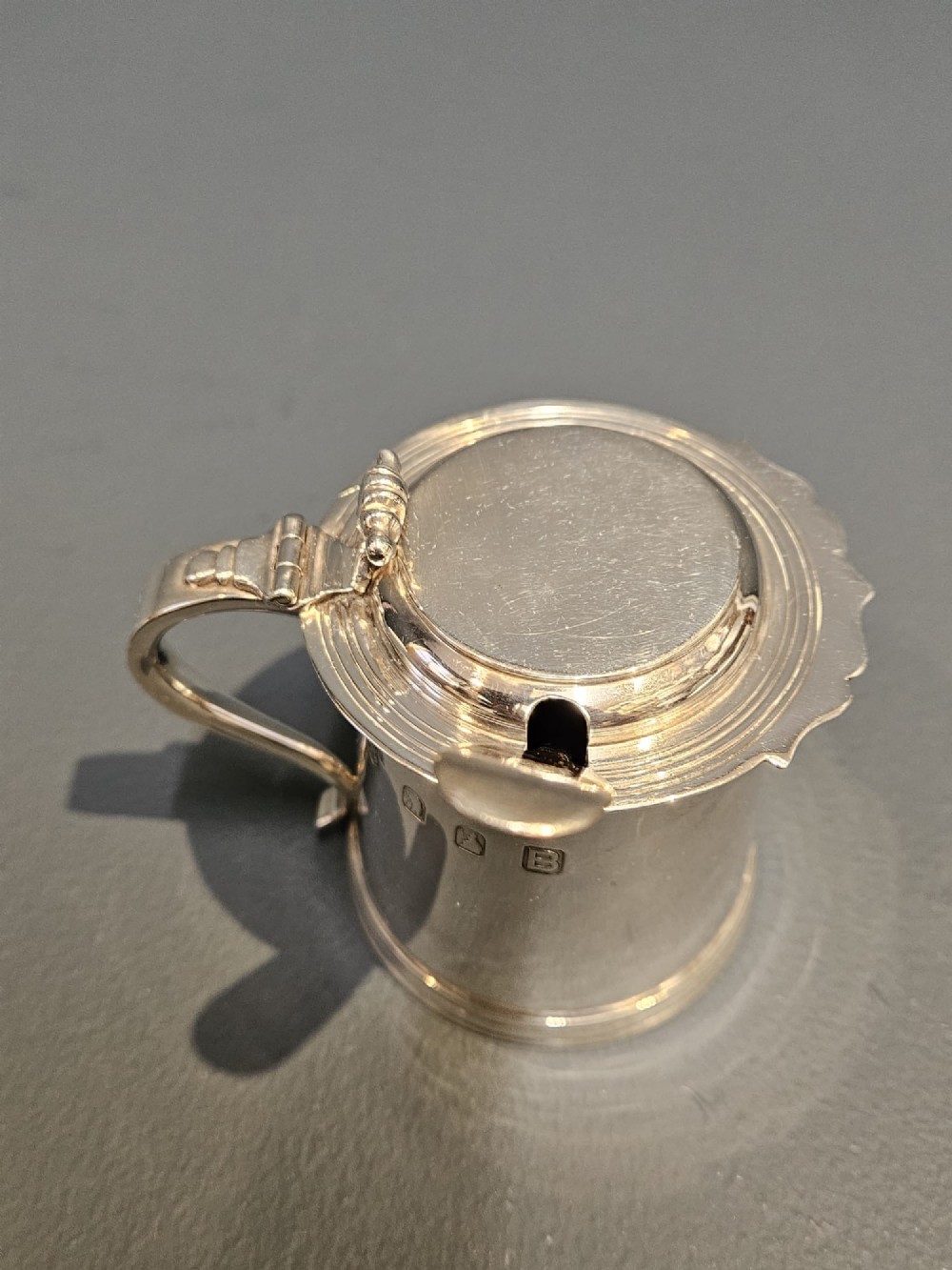 A cylindrical Britannia silver mustard pot featuring a scalloped collar rim, stepped lid with shell finial, and an inserted silver spoon. View II
