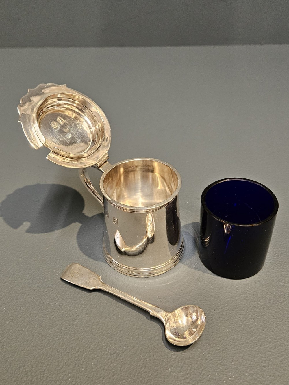 A cylindrical Britannia silver mustard pot featuring a scalloped collar rim, stepped lid with shell finial, and an inserted silver spoon. View V