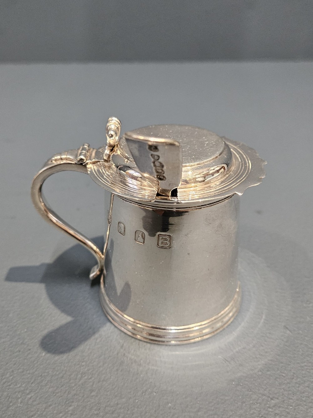 A cylindrical Britannia silver mustard pot featuring a scalloped collar rim, stepped lid with shell finial, and an inserted silver spoon. View VII