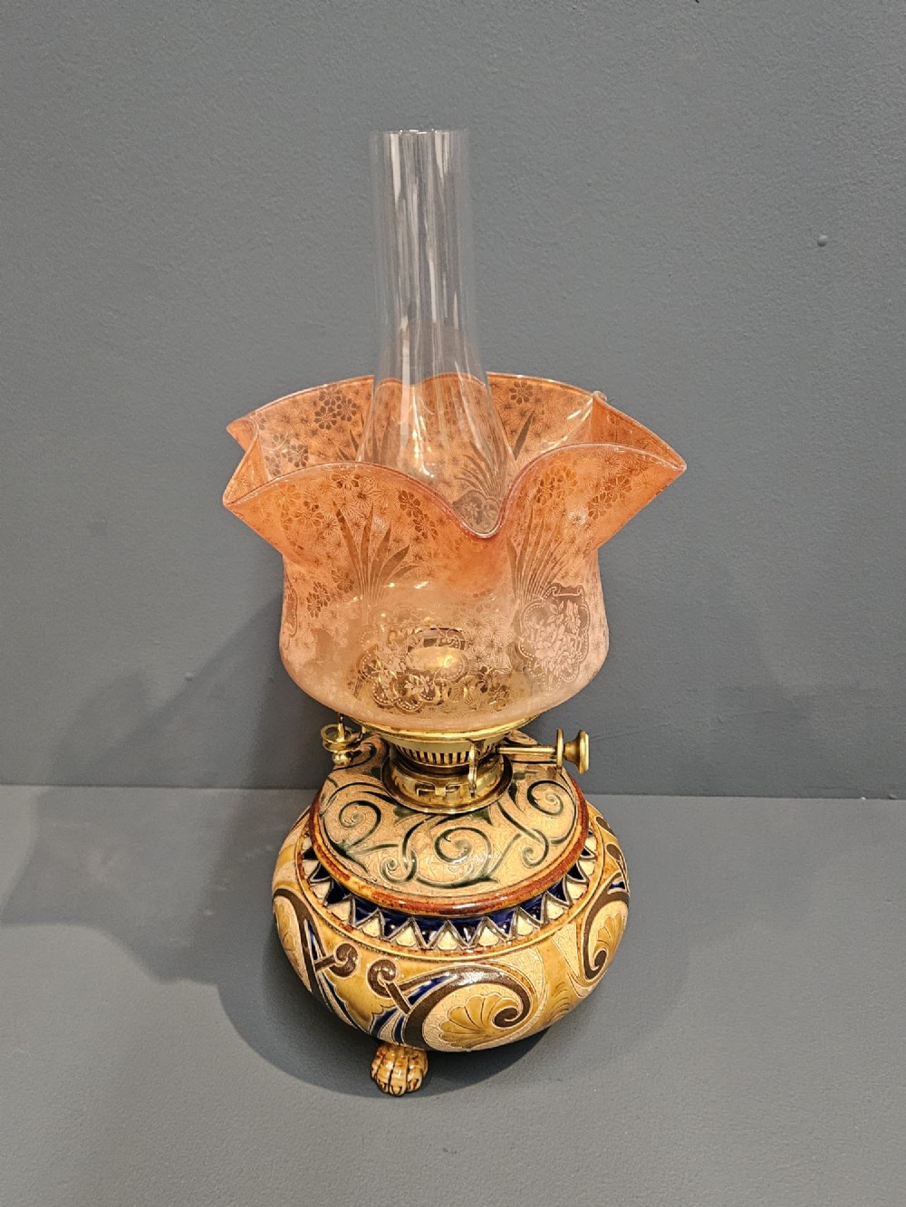 A Victorian Doulton Lambeth stoneware oil lamp featuring an incised ceramic base on claw feet, a brass burner, a glass chimney, and a frilled pink etched shade. View VI
