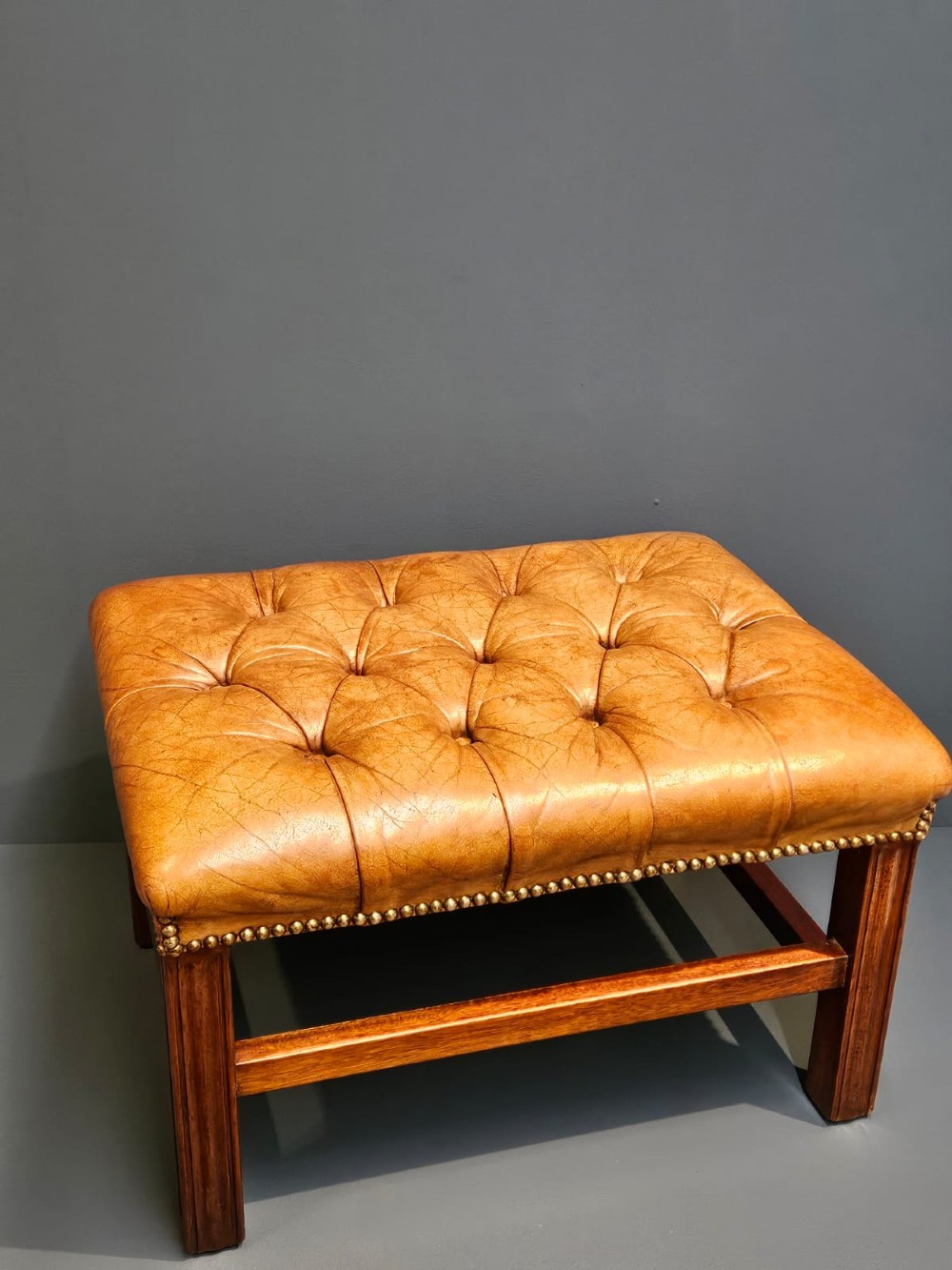 A rectangular 1930s footstool featuring deep-buttoned tan leather upholstery, brass studding along the perimeter, and square timber legs with stretchers.
