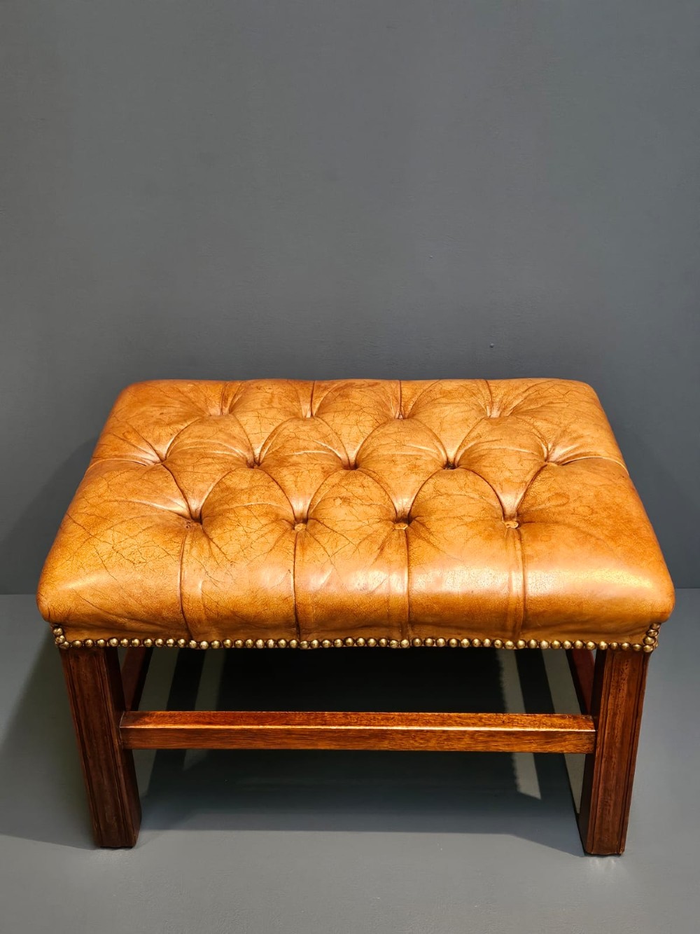 A rectangular 1930s footstool featuring deep-buttoned tan leather upholstery, brass studding along the perimeter, and square timber legs with stretchers. View II