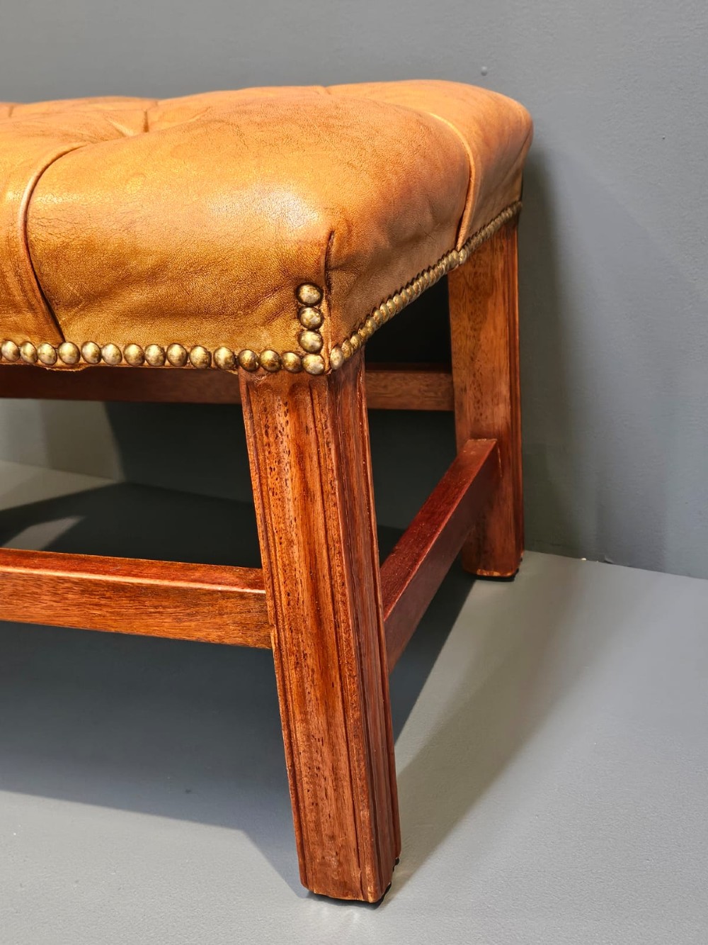 A rectangular 1930s footstool featuring deep-buttoned tan leather upholstery, brass studding along the perimeter, and square timber legs with stretchers. View IV