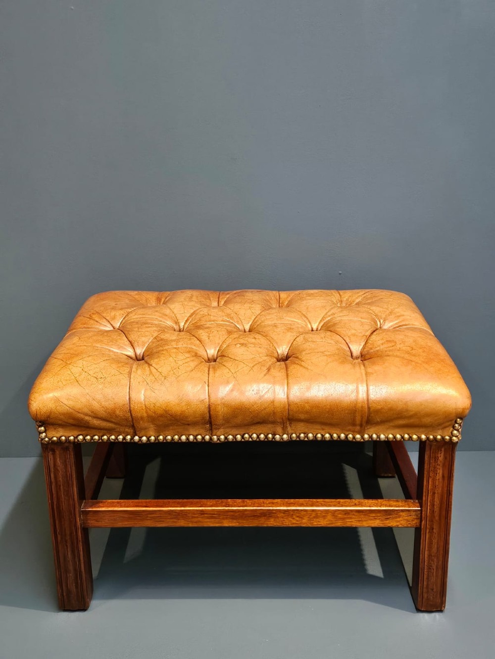 A rectangular 1930s footstool featuring deep-buttoned tan leather upholstery, brass studding along the perimeter, and square timber legs with stretchers. View V