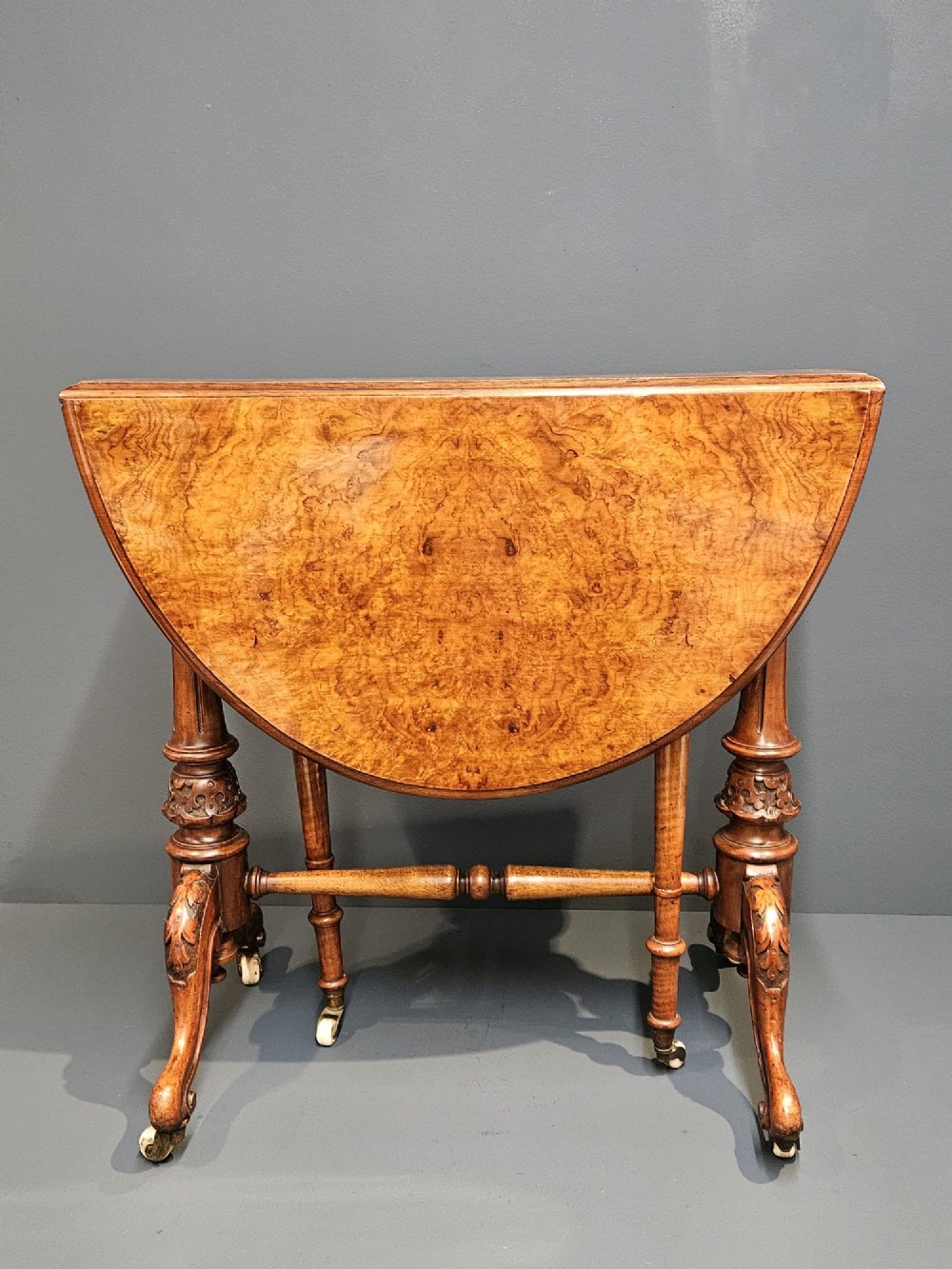 A Victorian burr walnut baby Sutherland table featuring two drop leaves, dual turned column supports, and carved feet on white ceramic castors. View III