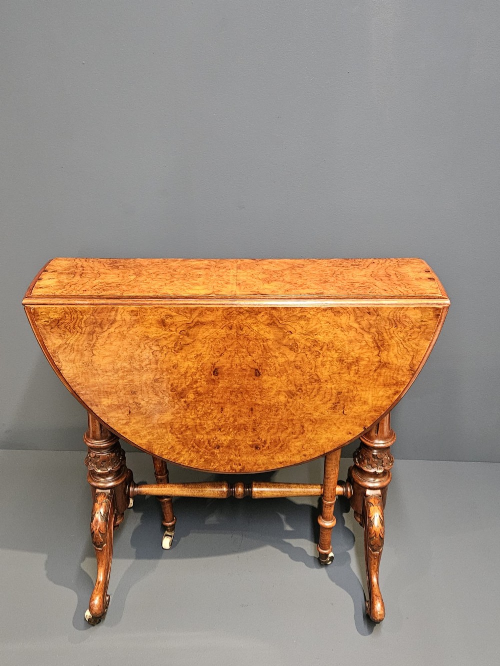 A Victorian burr walnut baby Sutherland table featuring two drop leaves, dual turned column supports, and carved feet on white ceramic castors. View IV
