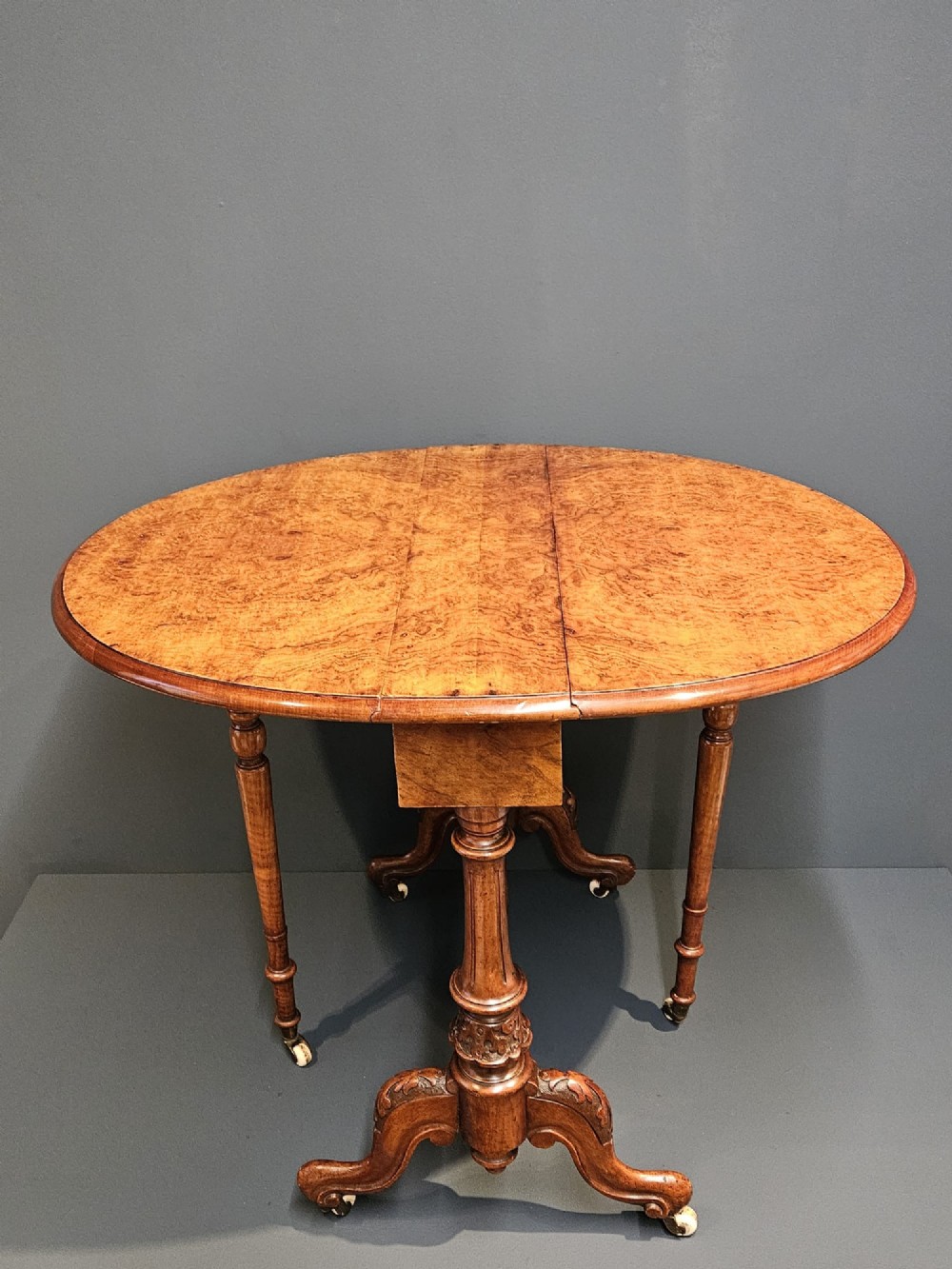 A Victorian burr walnut baby Sutherland table featuring two drop leaves, dual turned column supports, and carved feet on white ceramic castors. View V