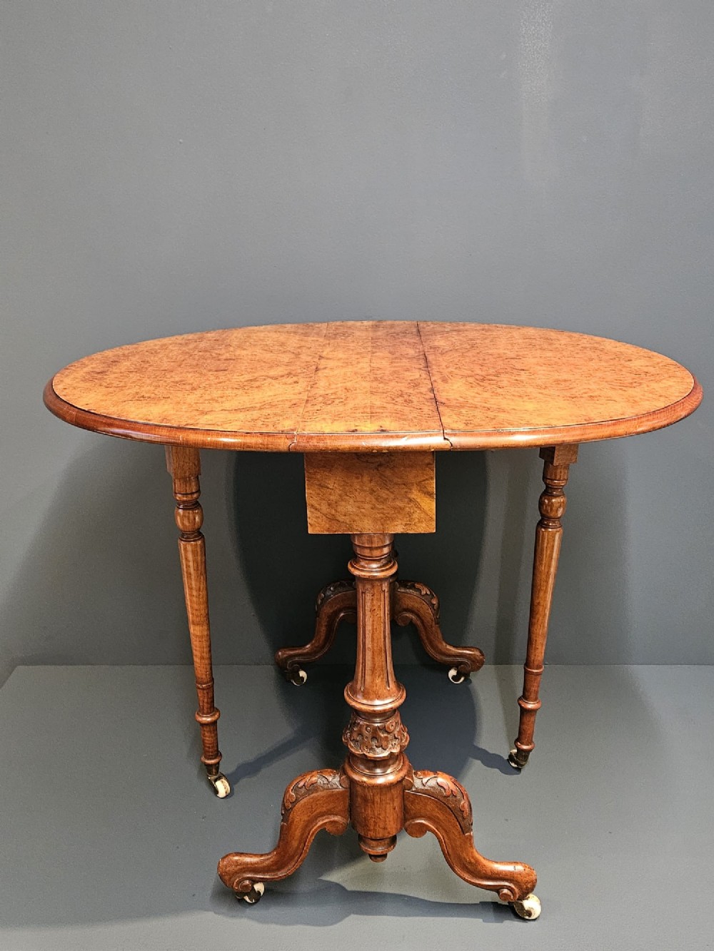 A Victorian burr walnut baby Sutherland table featuring two drop leaves, dual turned column supports, and carved feet on white ceramic castors. View IX