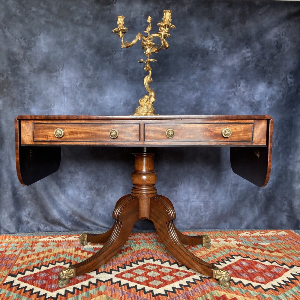A Regency mahogany sofa table featuring two drop-leaf ends, twin frieze drawers with brass handles, ebony inlay, and reeded sabre legs with brass paw castors. View V