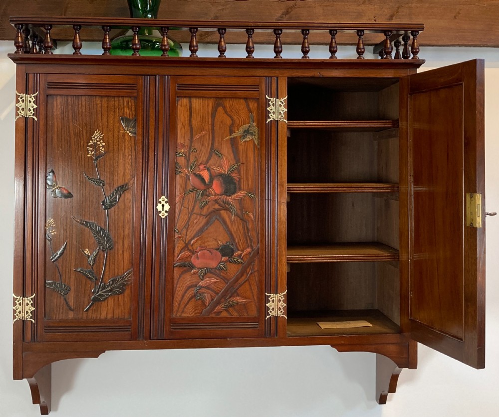 An Aesthetic Movement walnut hanging cabinet featuring three front doors decorated with carved nature motifs, a mirrored panel, exposed brass hinges, and a spindled top gallery. View III