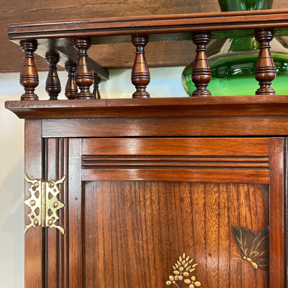 An Aesthetic Movement walnut hanging cabinet featuring three front doors decorated with carved nature motifs, a mirrored panel, exposed brass hinges, and a spindled top gallery. View V