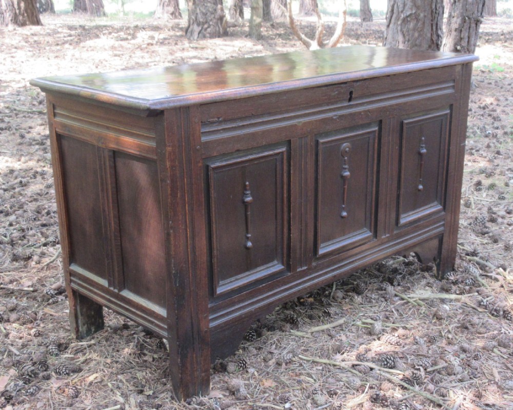 A mid-17th-century dark oak coffer featuring a flat hinged lid, triple-panelled front with split-baluster detail, and sturdy stile legs.