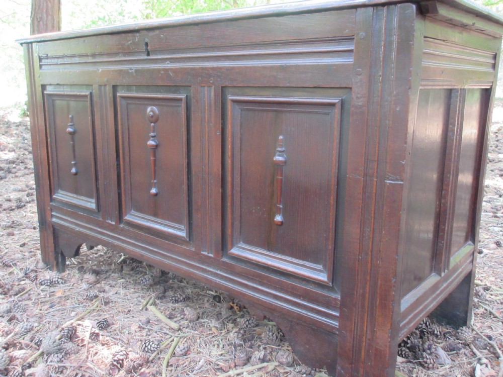 A mid-17th-century dark oak coffer featuring a flat hinged lid, triple-panelled front with split-baluster detail, and sturdy stile legs. View II