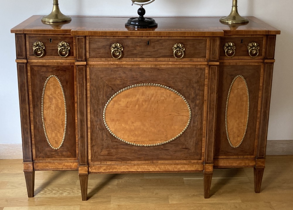 A late 18th-century George III breakfront wooden side cabinet featuring central and flanking drawers above doors decorated with oval satinwood marquetry panels and brass ring handles.