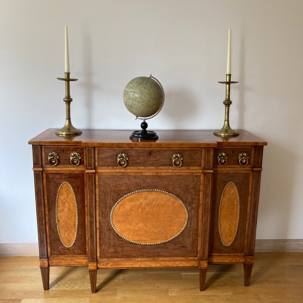 A late 18th-century George III breakfront wooden side cabinet featuring central and flanking drawers above doors decorated with oval satinwood marquetry panels and brass ring handles. View II