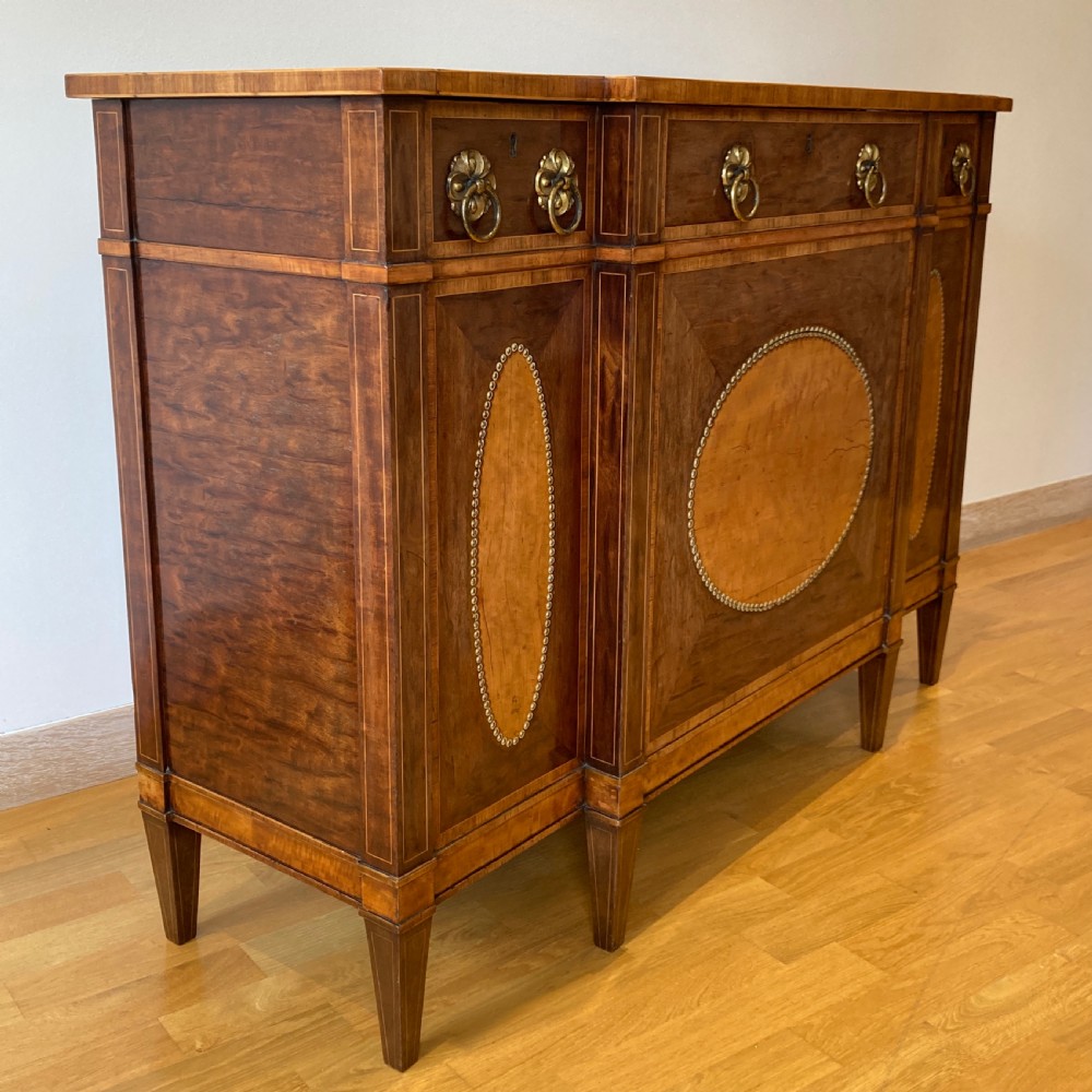 A late 18th-century George III breakfront wooden side cabinet featuring central and flanking drawers above doors decorated with oval satinwood marquetry panels and brass ring handles. View III