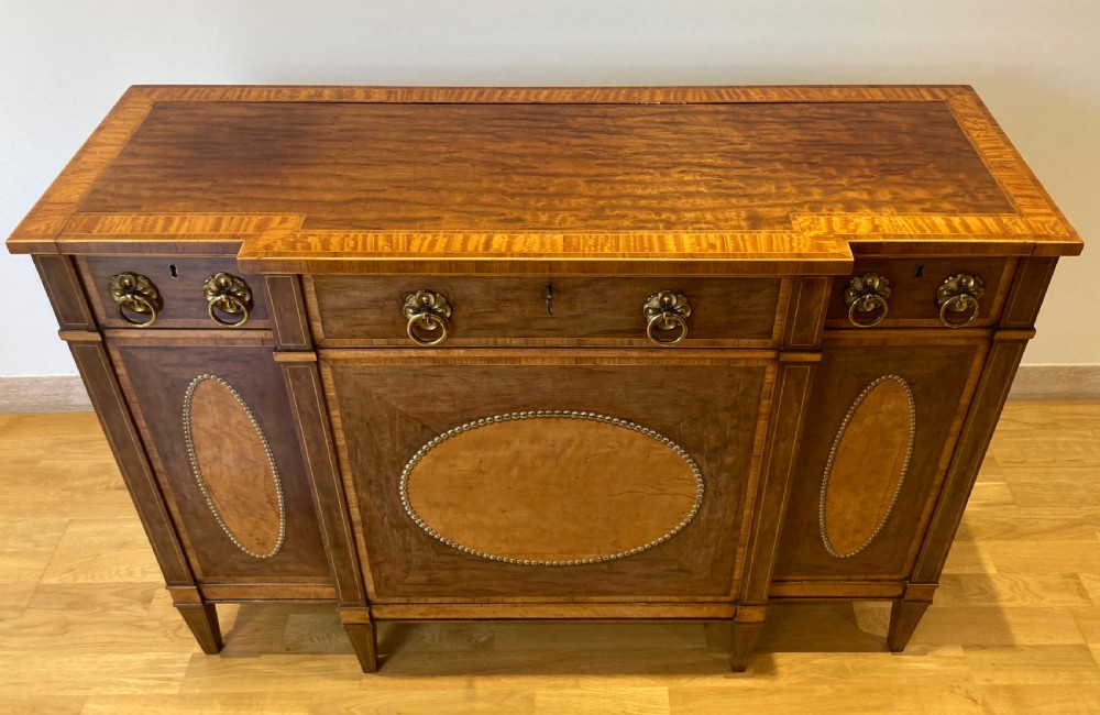 A late 18th-century George III breakfront wooden side cabinet featuring central and flanking drawers above doors decorated with oval satinwood marquetry panels and brass ring handles. View V