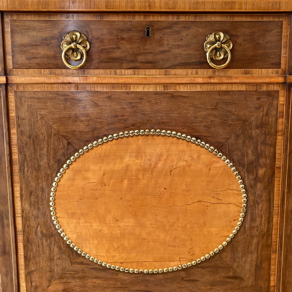 A late 18th-century George III breakfront wooden side cabinet featuring central and flanking drawers above doors decorated with oval satinwood marquetry panels and brass ring handles. View VII