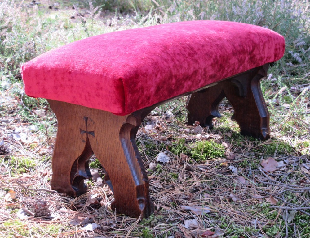 A Victorian Gothic Revival dark oak footstool featuring carved trestle supports with an incised cross, upholstered in red velvet fabric.