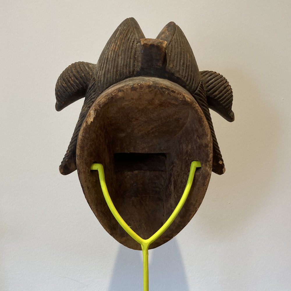 A carved wooden Punu tribal mask featuring a white kaolin face, arched eyebrows, narrow slit eyes, dark lips, and an elaborate dark coiffure, mounted on a yellow stand. View VIII