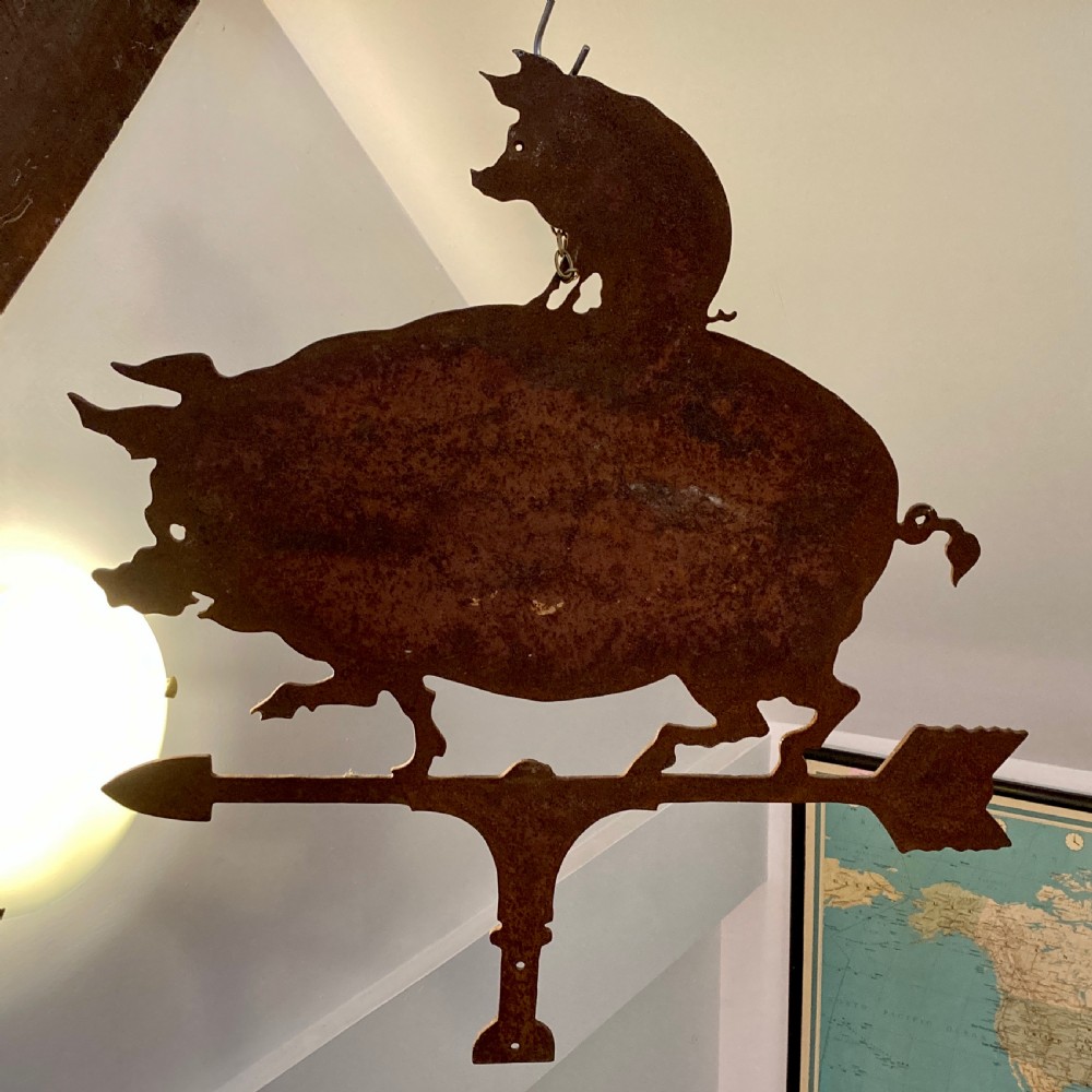 A wrought iron weather vane featuring a silhouette of a large pig with a smaller pig on its back, mounted on a directional arrow with a weathered brown patina.