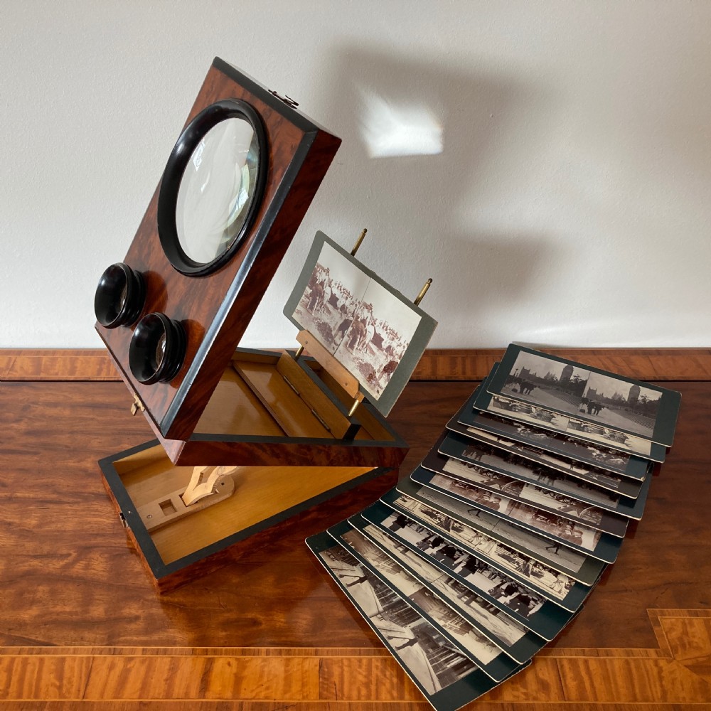 An antique Victorian hinged burr yew wood tabletop stereoscope shown open, featuring dual circular viewing lenses, a central magnifying lens, and a selection of stereoview cards.