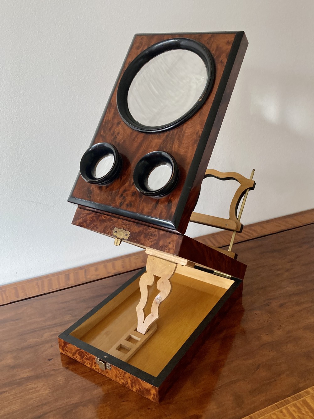 An antique Victorian hinged burr yew wood tabletop stereoscope shown open, featuring dual circular viewing lenses, a central magnifying lens, and a selection of stereoview cards. View V