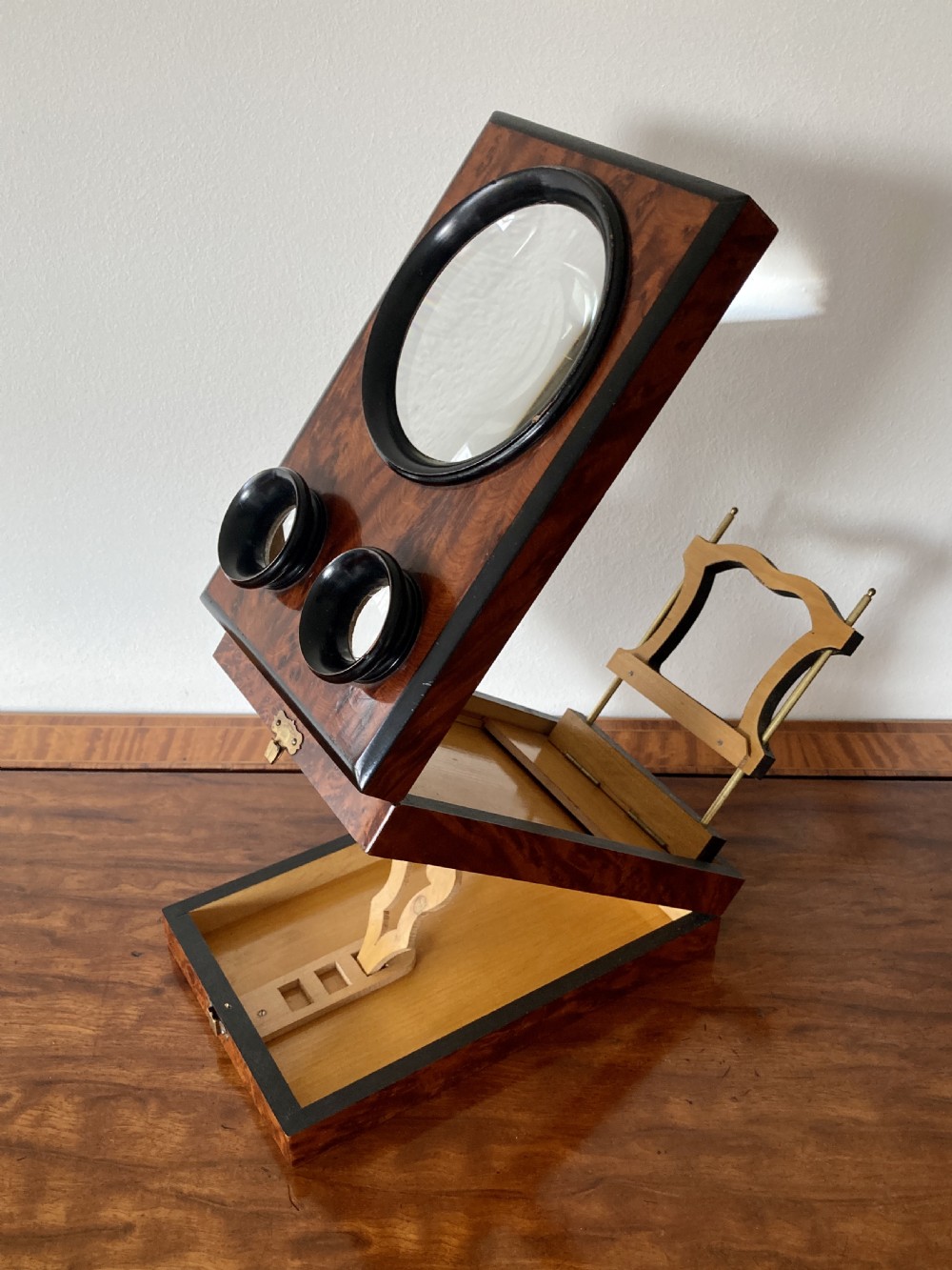 An antique Victorian hinged burr yew wood tabletop stereoscope shown open, featuring dual circular viewing lenses, a central magnifying lens, and a selection of stereoview cards. View VI