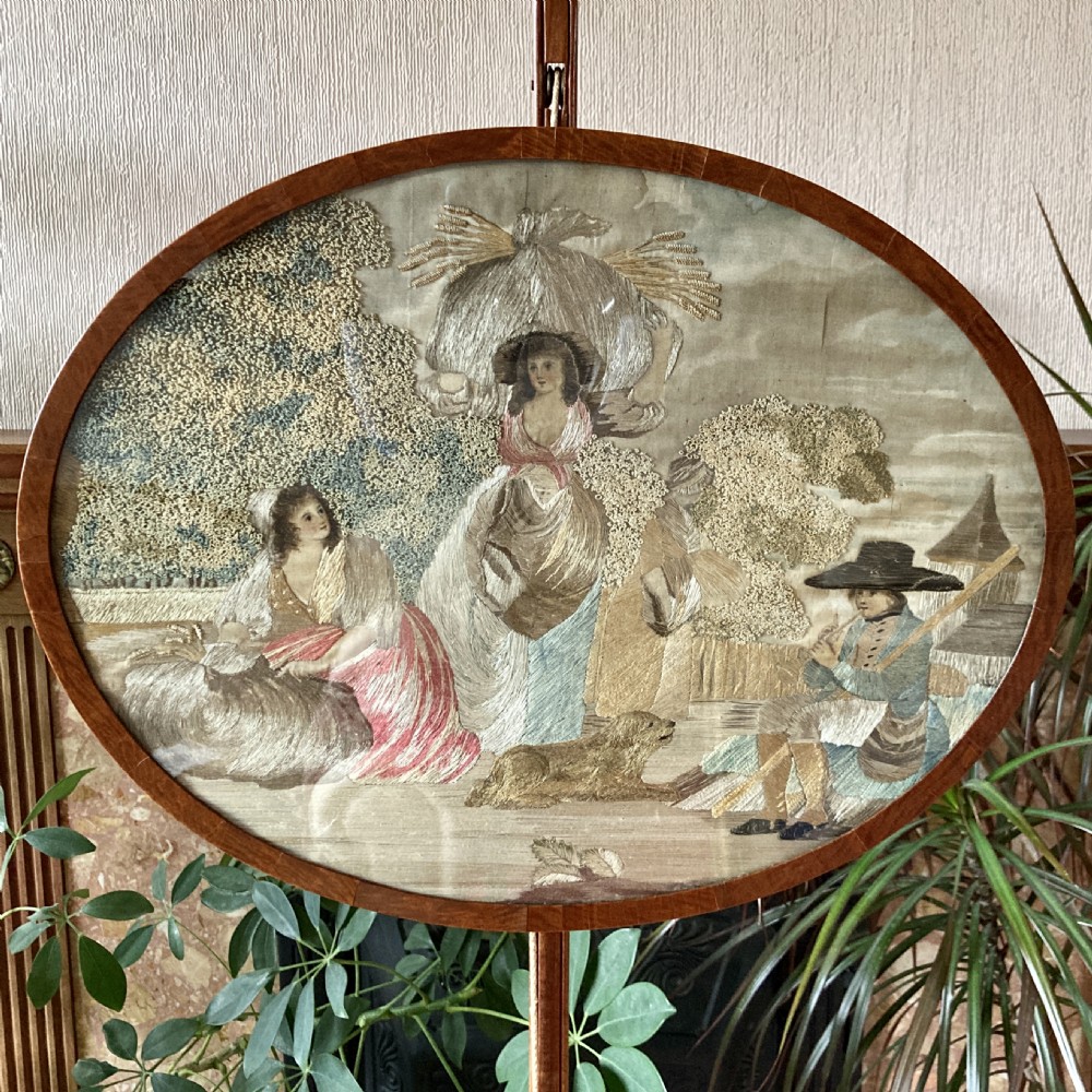 An oval mahogany fire screen containing an 18th-century silk needlework scene of pastoral figures, mounted on a central wooden pole.