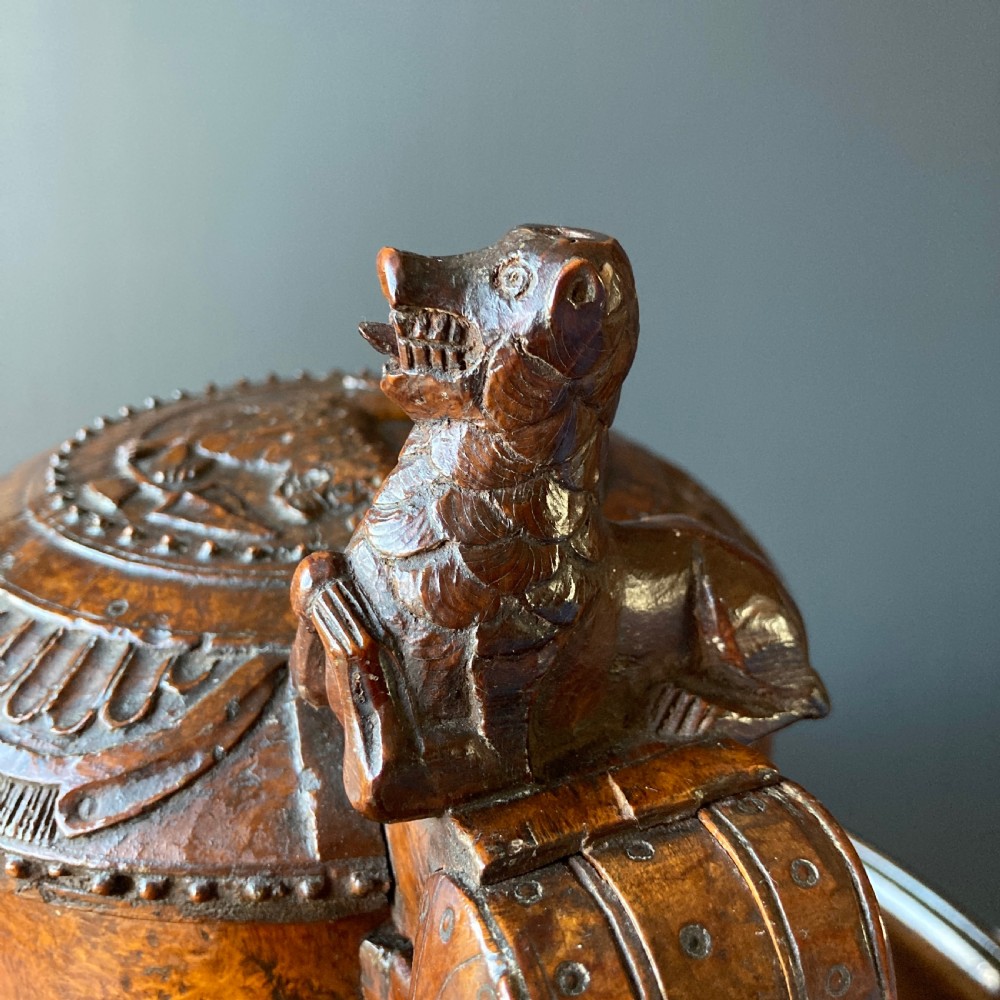 An 18th-century Norwegian carved wooden peg tankard featuring a lion thumbpiece, domed relief-carved lid, and carved lion feet in a warm brown finish. View X