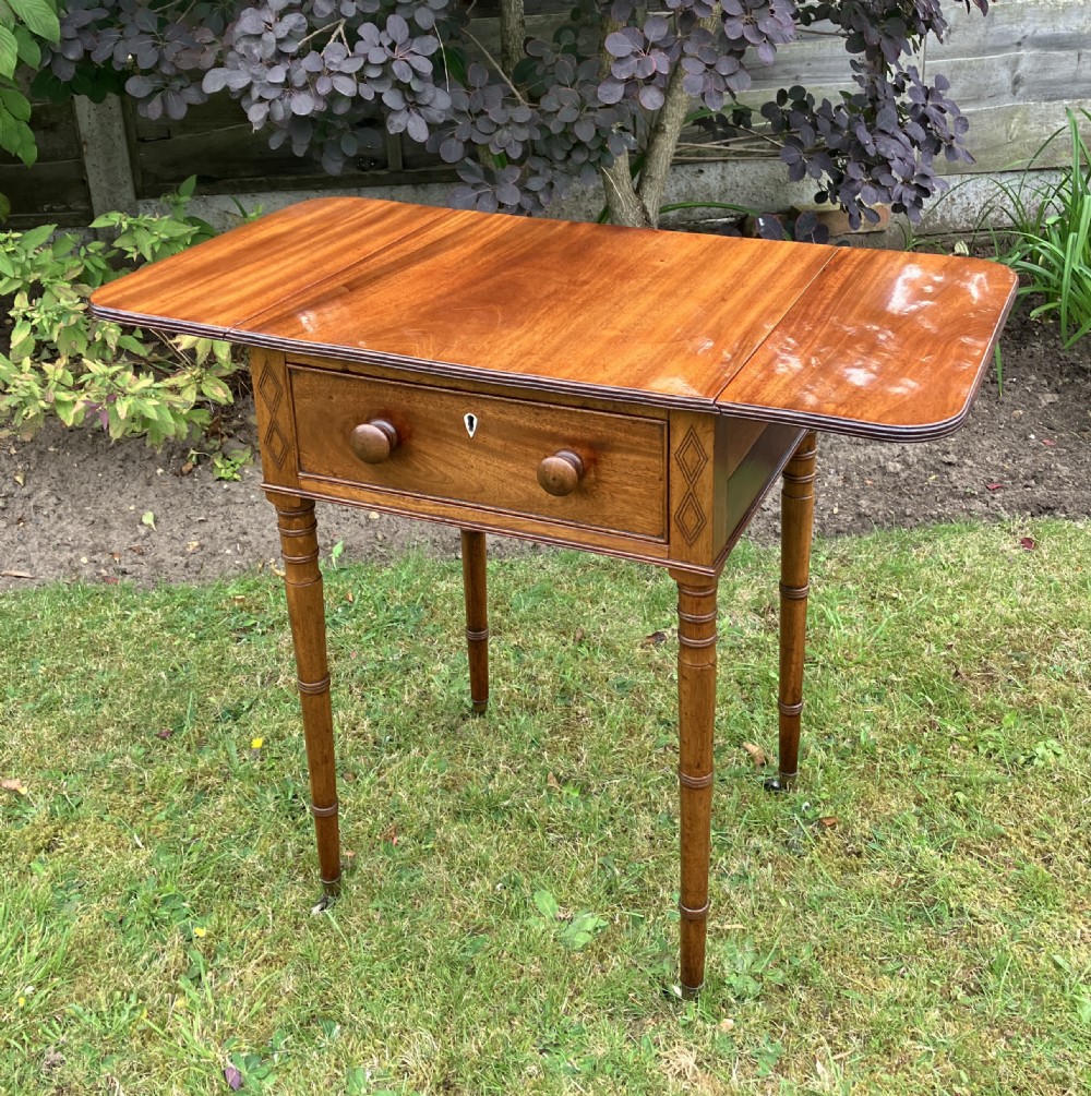 A late Georgian mahogany work table featuring twin drop leaves, a single drawer with wooden knobs, inlaid diamond details, and turned legs on brass castors. View V